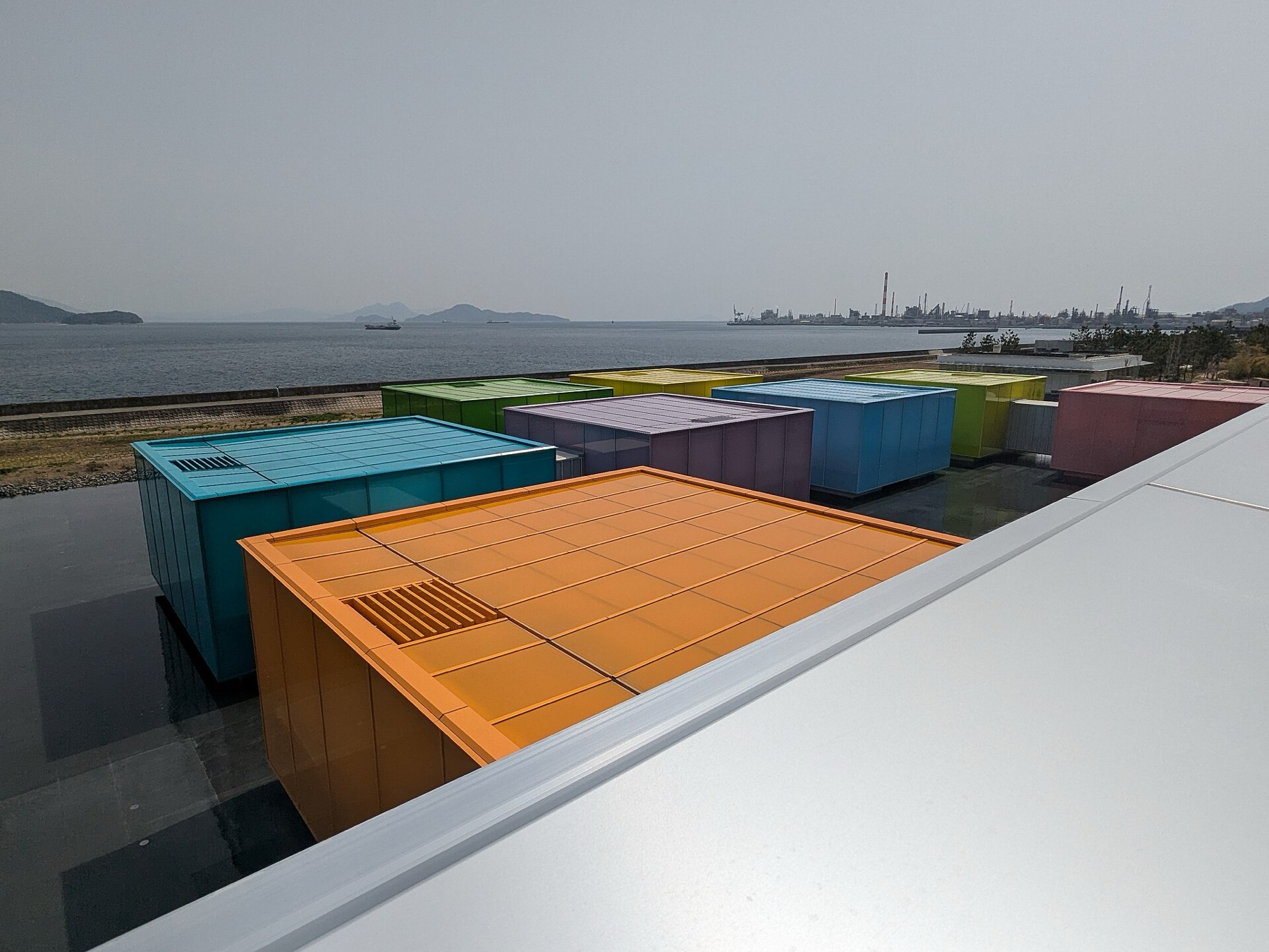 Eight brightly coloured movable gallery boxes in orange, teal, purple, green, yellow and pink float on a shallow reflecting water basin at the Simose Art Museum in Otake, Hiroshima, with the Seto Inland Sea and distant islands behind them.
