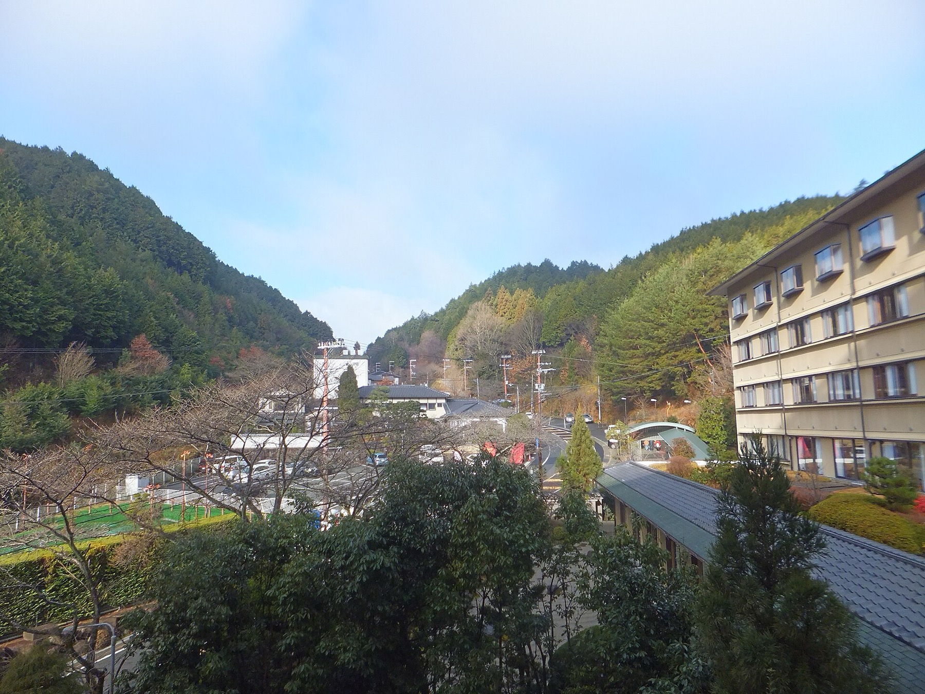 The Yunohana Onsen hot-spring valley near Kameoka in Kyoto Prefecture, showing forested hillsides in late autumn colour, a multi-storey ryokan building on the right, tiled roofs, a car park and a winding road running up the valley.