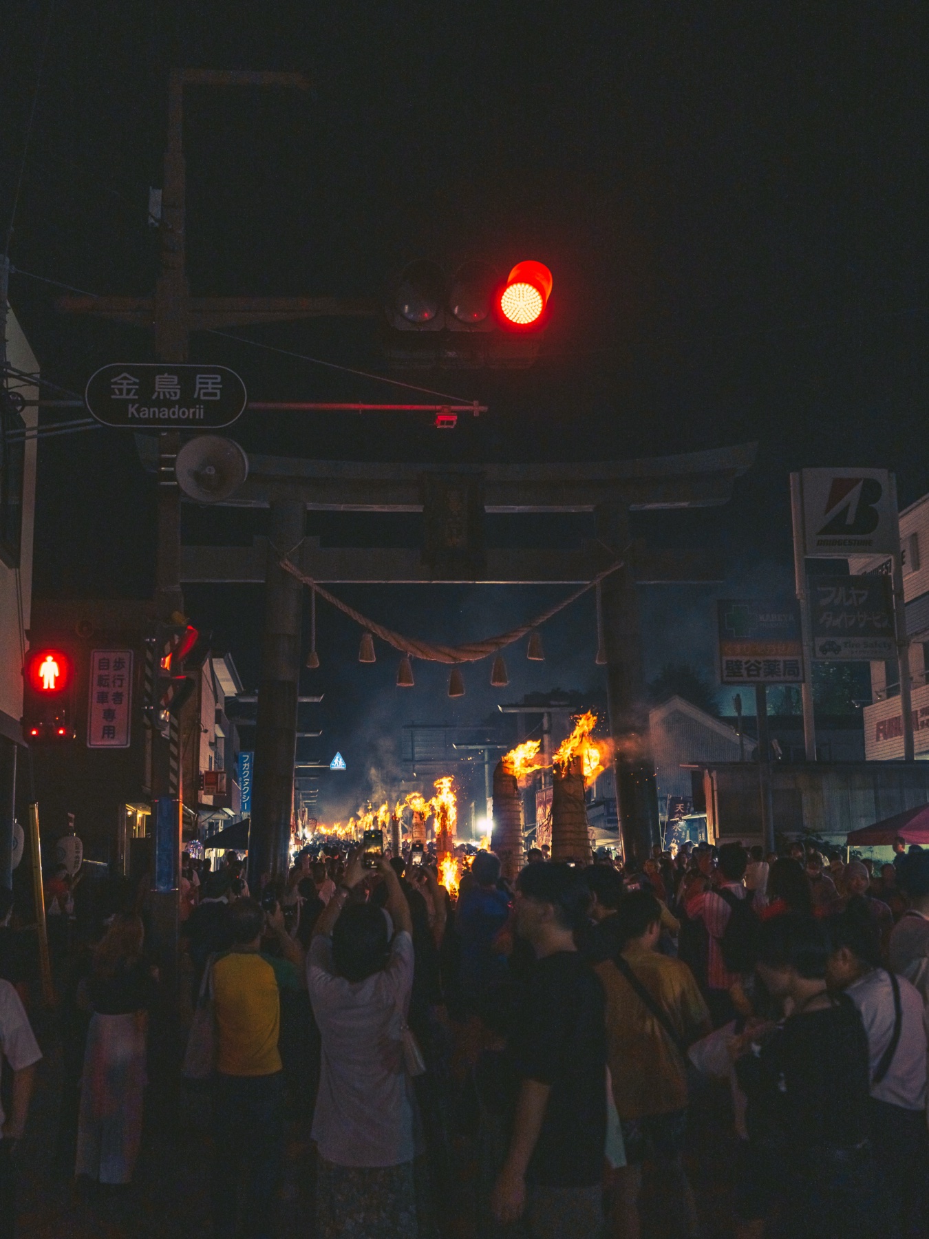 Festival crowds fill the street beneath the Kanadorii gate sign and a red traffic light at night, with taimatsu torch fires burning down the closed main street of Fujiyoshida toward Mt Fuji.