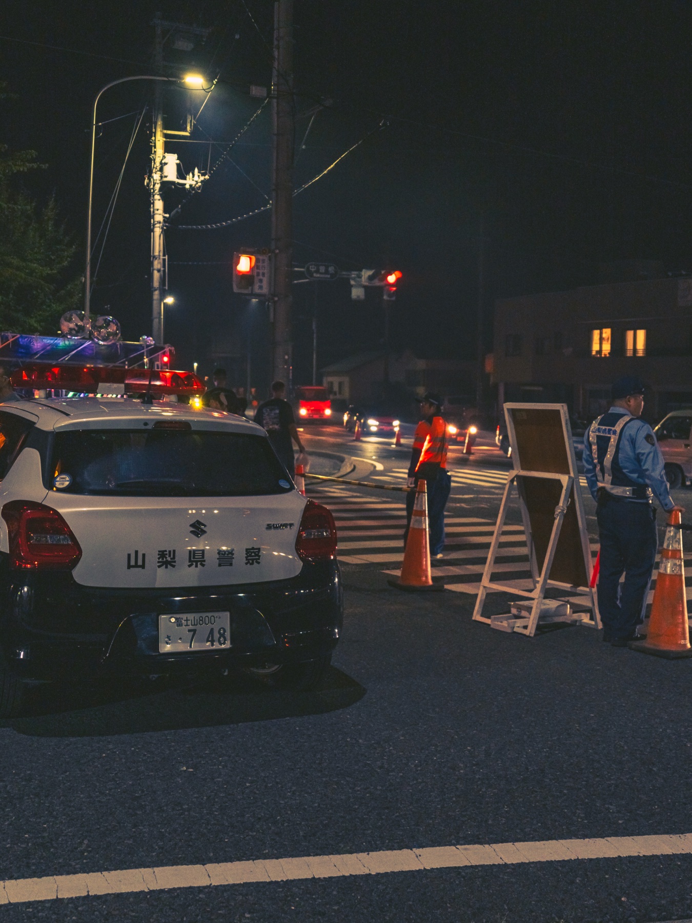 A Yamanashi prefectural police car, traffic cones and officers block a junction in Fujiyoshida at night for the Yoshida Fire Festival road closures, with traffic being diverted away from the festival streets.