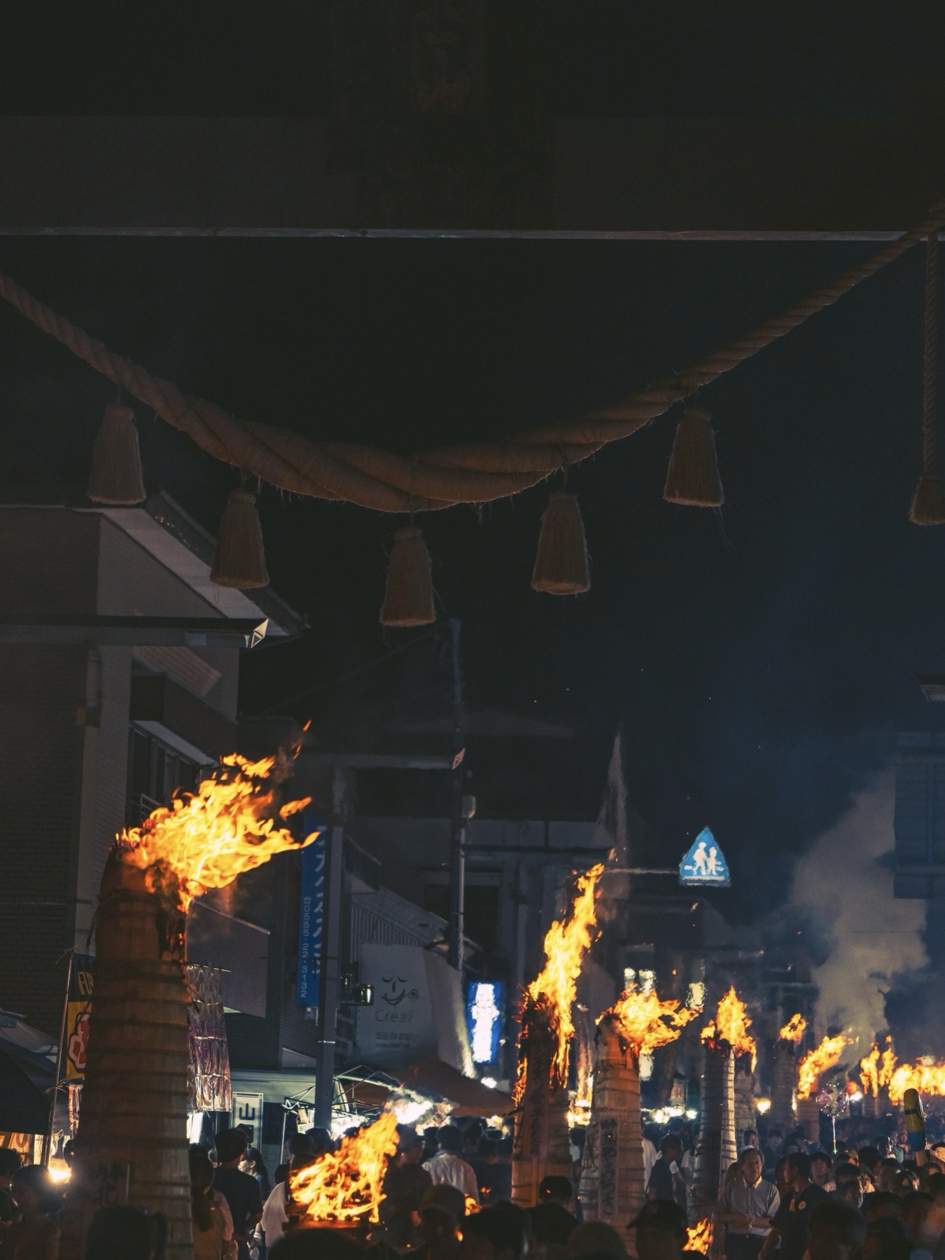 The sacred straw shimenawa rope with hanging tassels crosses the frame above a receding line of burning taimatsu torches and festival crowds on the main street of Fujiyoshida at night.