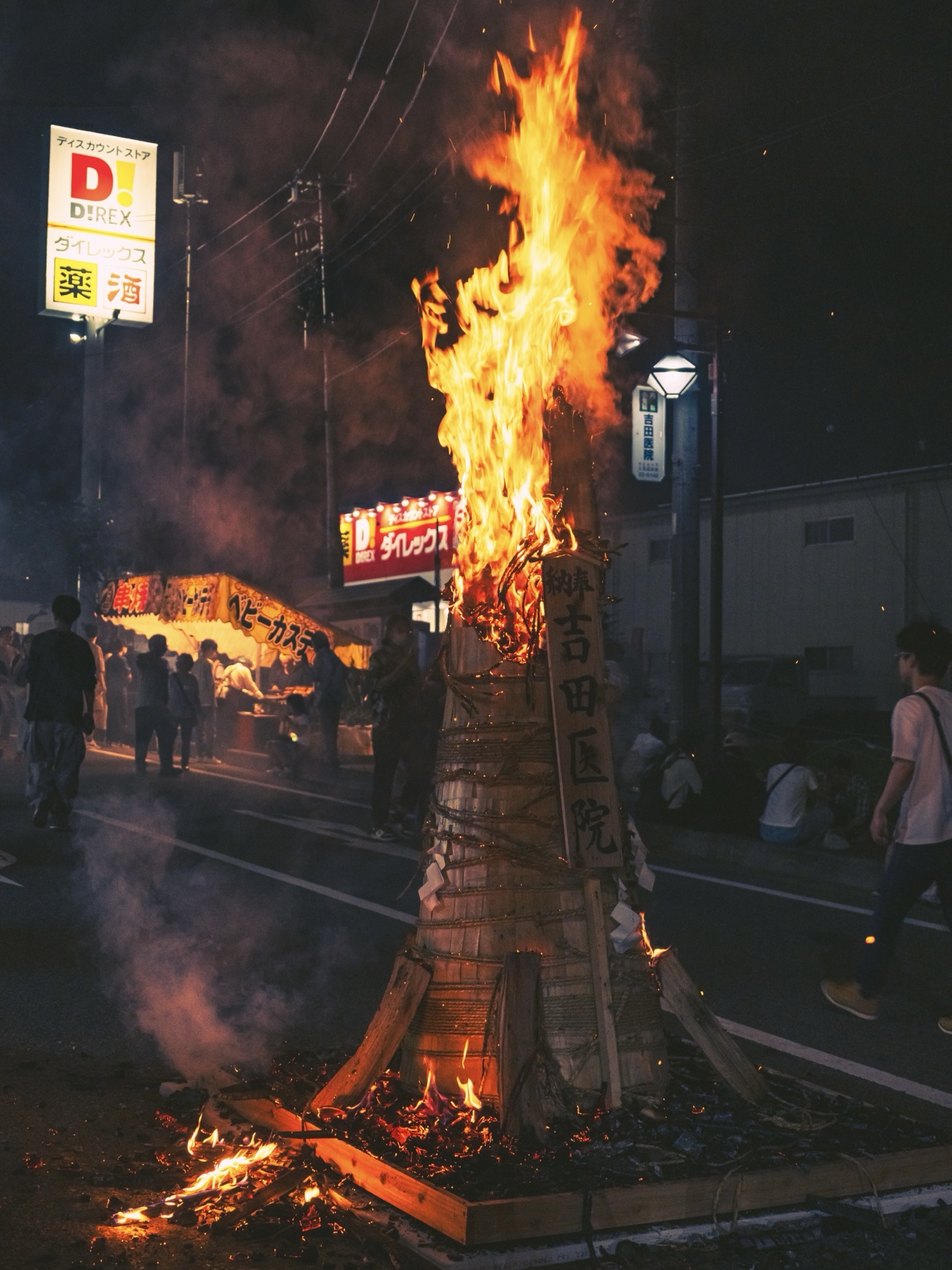 A close-up of a three-metre taimatsu torch in full blaze at the Yoshida Fire Festival, with a wooden offering plaque bearing a local clinic's name wired to its side and food stalls lit behind.