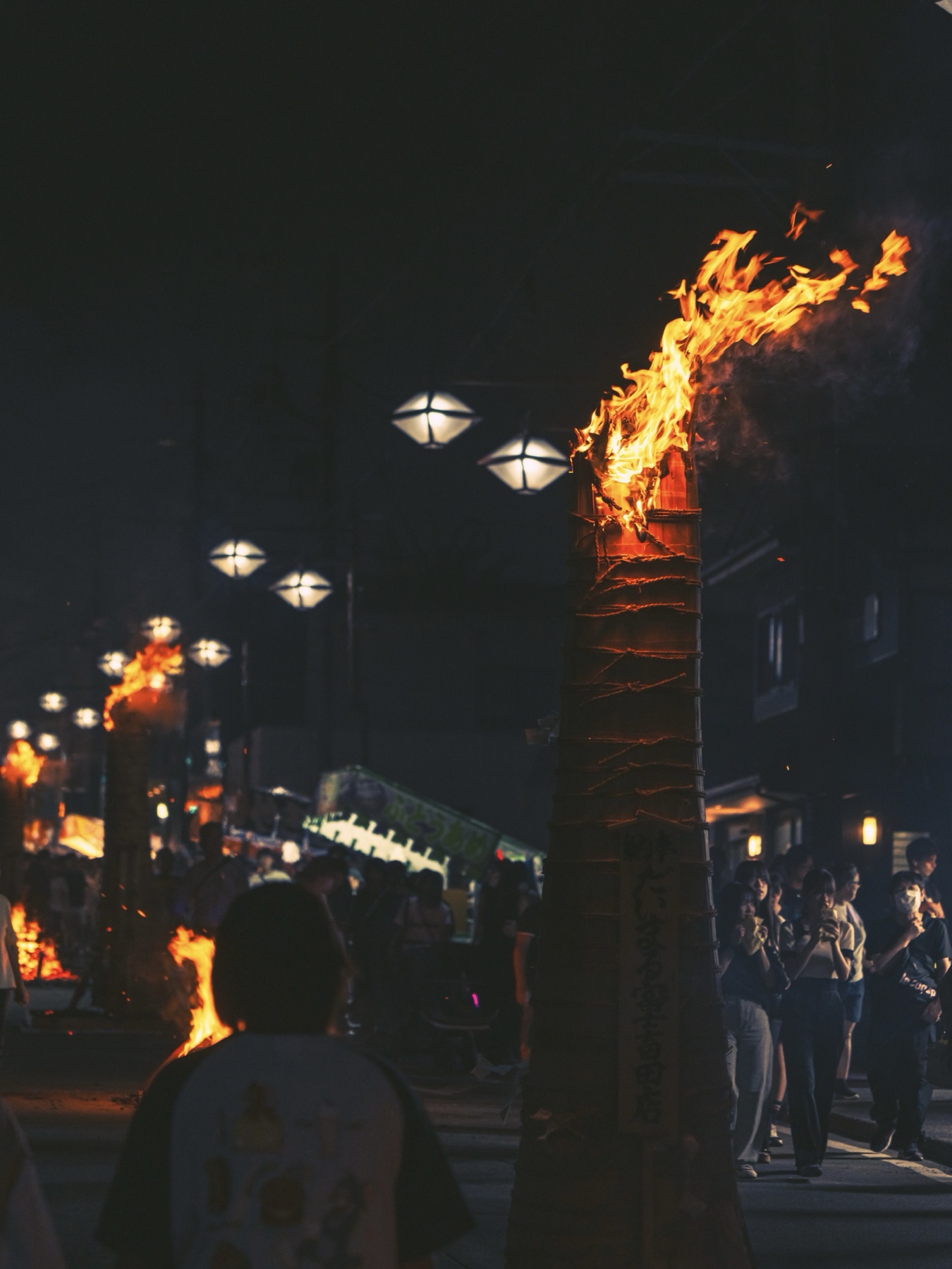 A single bamboo-shoot-shaped taimatsu torch burns fiercely in the foreground while diamond-shaped street lanterns and further torch fires line Fujiyoshida's festival street into the night.