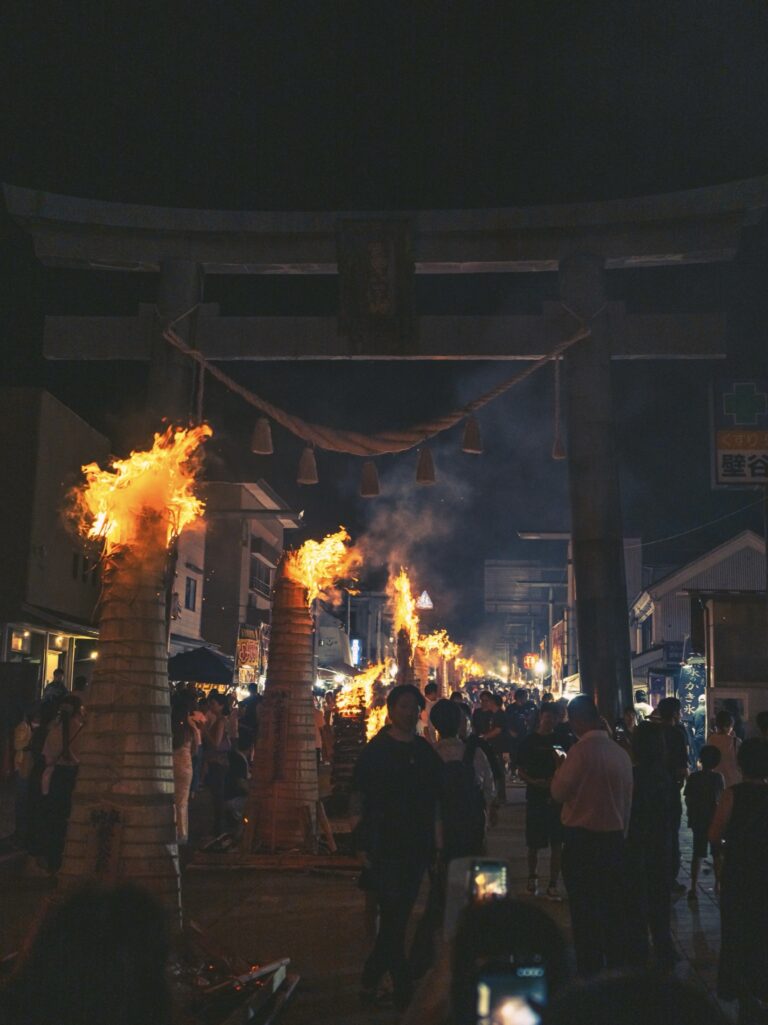 Rows of taimatsu torches blaze down Fujiyoshida main street at the Yoshida Fire Festival, framed by the great torii and its straw shimenawa rope, with crowds walking between the fires at dusk.