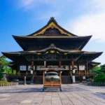 The National Treasure Main Hall of Zenkoji temple in Nagano City, a vast wooden structure rebuilt in 1707 with a sweeping hinoki-bark roof, seen from the front approach with worshippers on the steps
