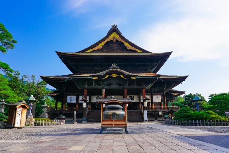 The National Treasure Main Hall of Zenkoji temple in Nagano City, a vast wooden structure rebuilt in 1707 with a sweeping hinoki-bark roof, seen from the front approach with worshippers on the steps