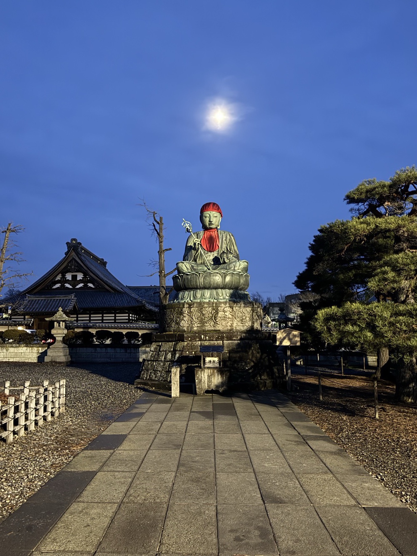 The seated bronze Nurebotoke Jizo statue of Zenkoji wearing a red hood beneath a bright moon in deep blue dawn light, with the tiled roofs of the temple precinct and a stone approach in the frame