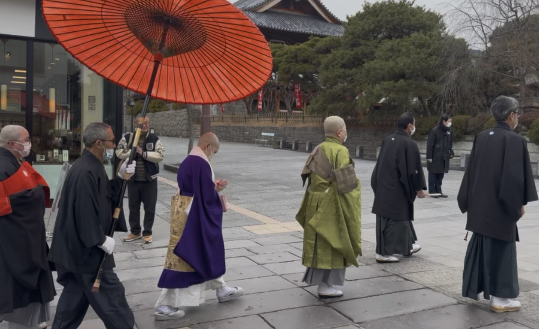 A procession of Buddhist priests in black, purple and green robes walking the stone approach of Zenkoji under a large red umbrella held by an attendant on a wet winter morning