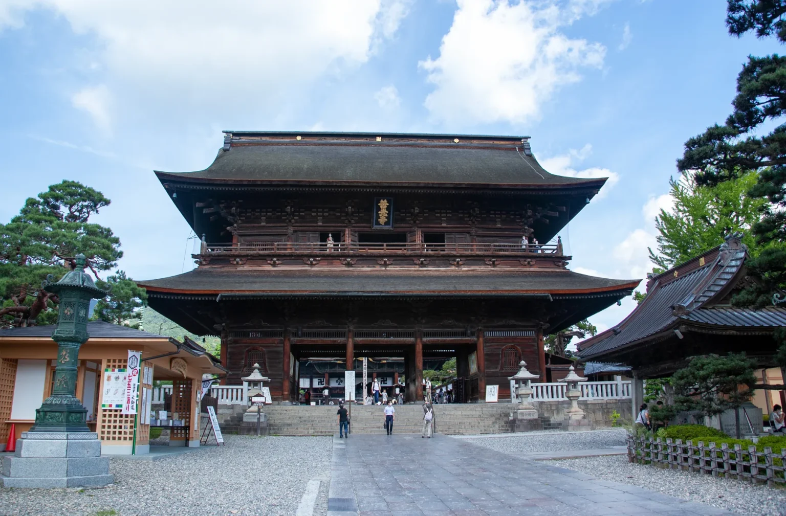 The two-storey wooden Sanmon gate of Zenkoji temple in Nagano with its dark cypress-bark roof, seen from the stone-paved approach with visitors passing beneath the central opening