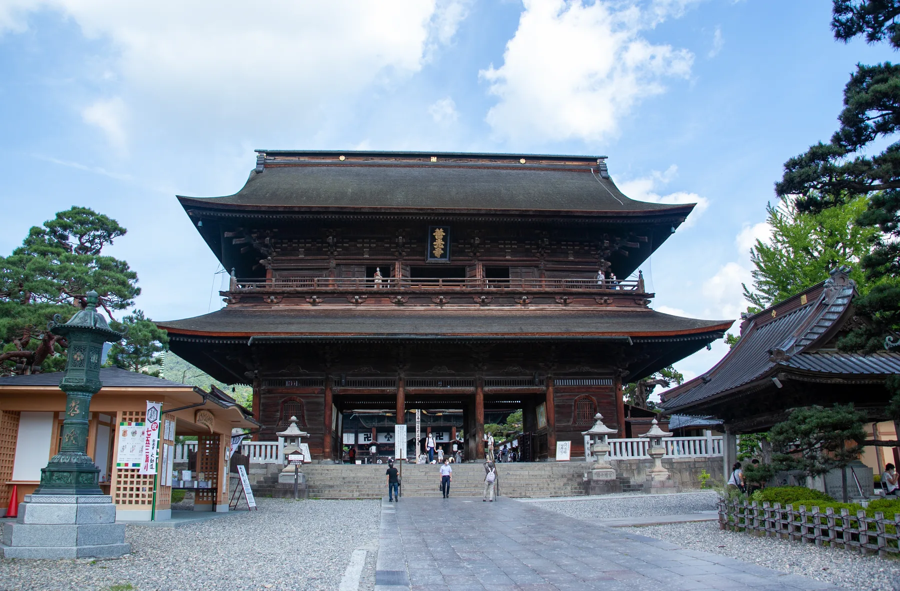 The two-storey wooden Sanmon gate of Zenkoji temple in Nagano with its dark cypress-bark roof, seen from the stone-paved approach with visitors passing beneath the central opening