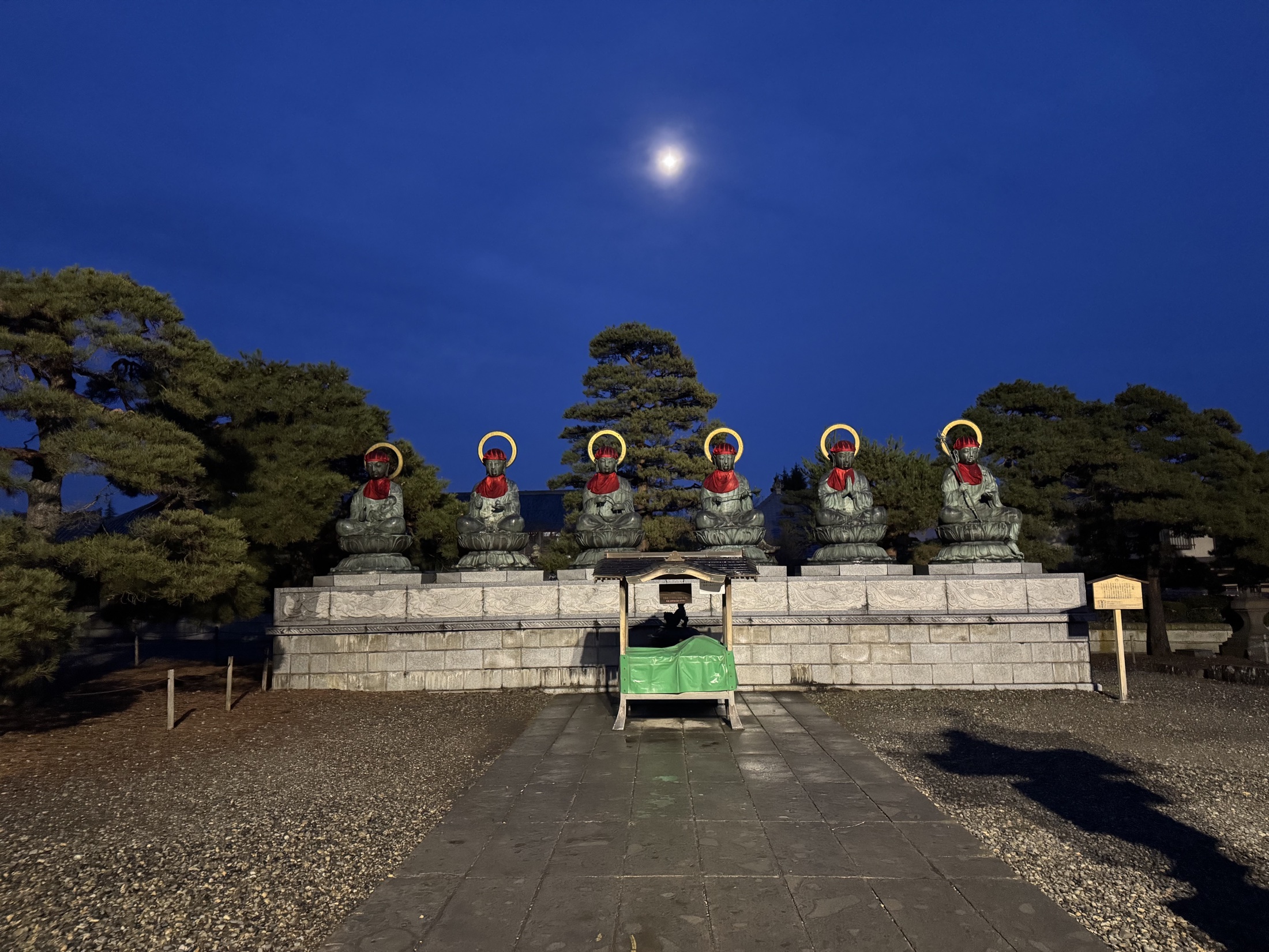 Six seated bronze Jizo statues with red bibs and golden halos at Zenkoji temple photographed in deep blue pre-dawn light with a bright moon overhead and pine trees behind them
