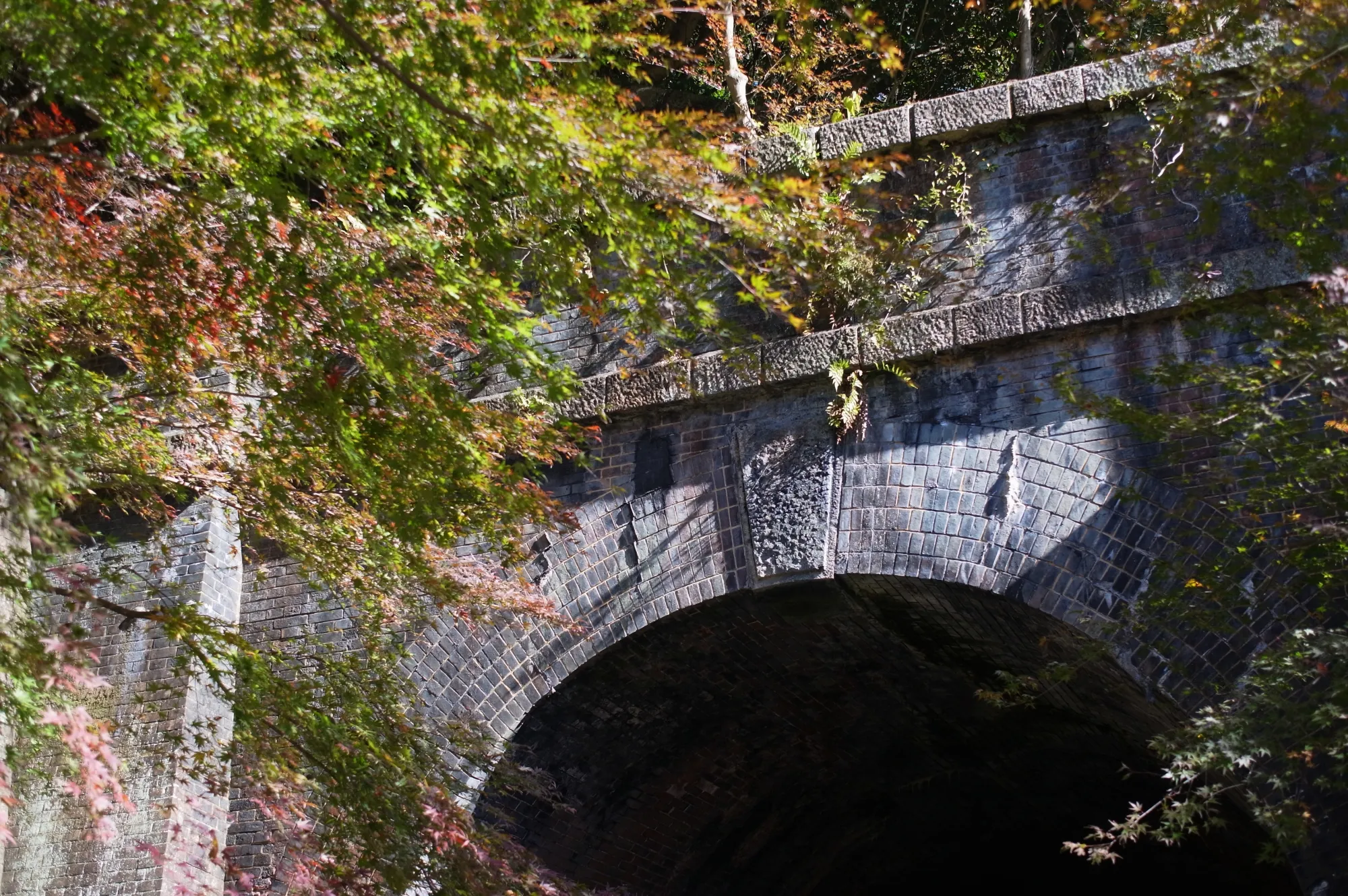 The arched portal of a Meiji-era railway tunnel in the Aigi Tunnel Group, built in brick with a rough-hewn stone keystone and a cut-stone coping course above the arch.