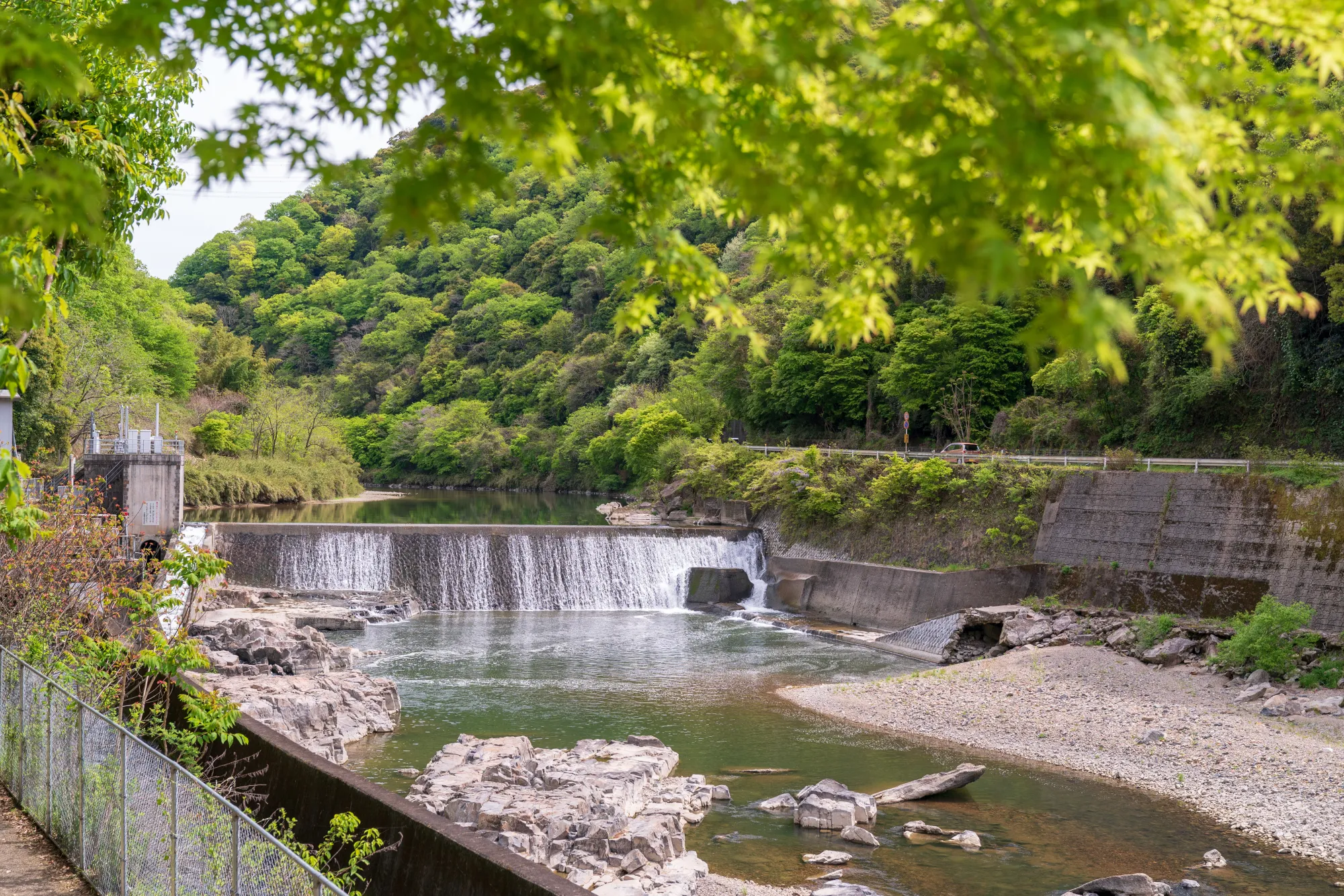 The Shonai River running through its gorge below the disused railway formation, with a low weir and wooded slopes in green summer foliage.
