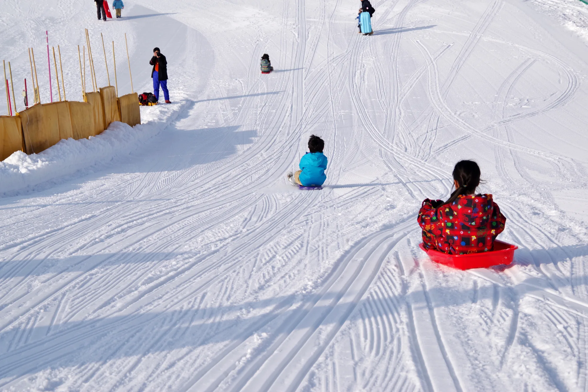 Two small children sledding down a gentle snow slope at a Japanese ski resort, one on a red plastic sled, with an orange safety fence along the edge and other families further up the hill