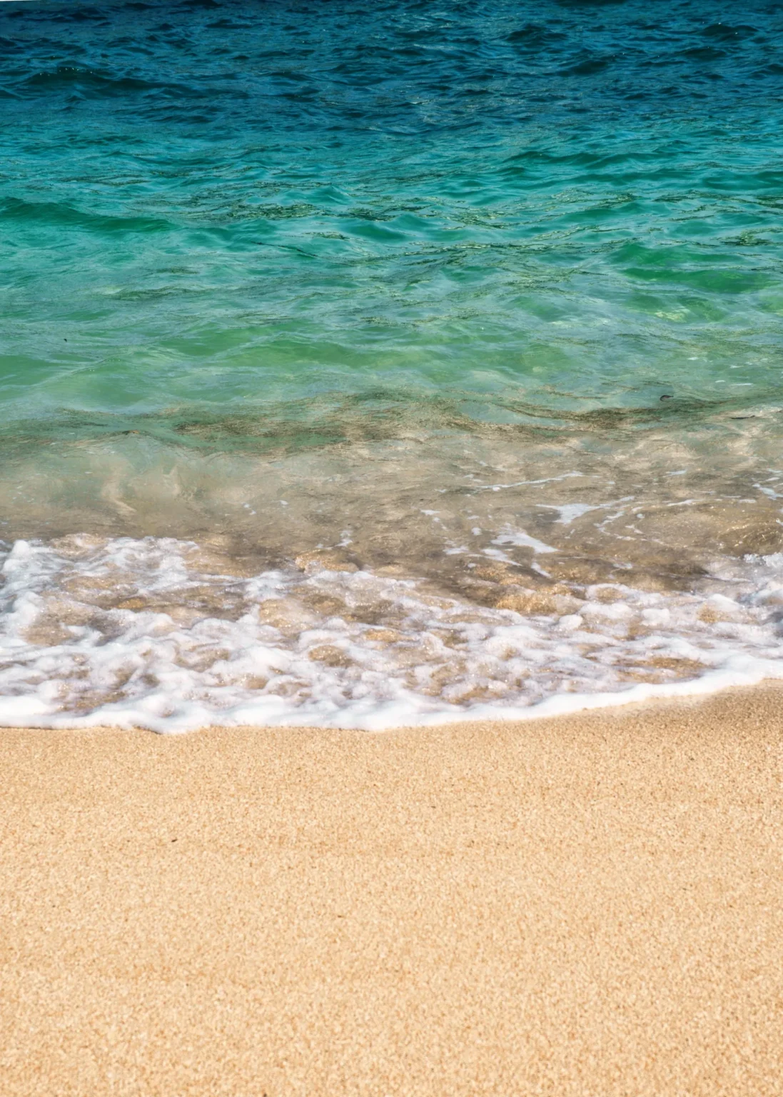 Clear turquoise water and a line of white foam washing onto a pale sandy beach in bright sunlight