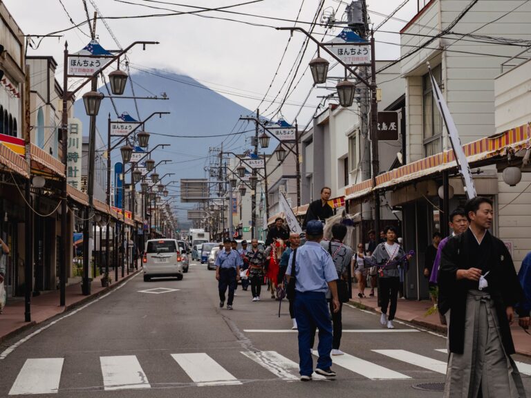 A white festival horse in purple regalia is led down Honcho-dori shopping street in Fujiyoshida during the September yabusame procession, with Mount Fuji rising directly over the retro shopfronts and street lamps
