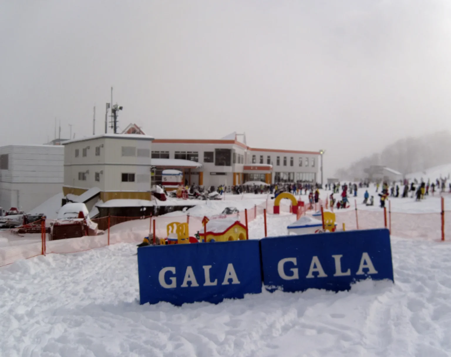 The Cheers mid-mountain building at GALA Yuzawa on an overcast day, with blue GALA banners in the snow in the foreground and crowds of skiers on the slope beside the children's play area.