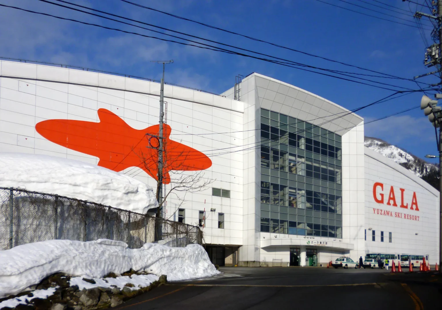 The white station building of GALA Yuzawa, with the resort name and a large orange logo on its wall, banks of cleared snow in front and a snowy ridge behind under a blue sky.