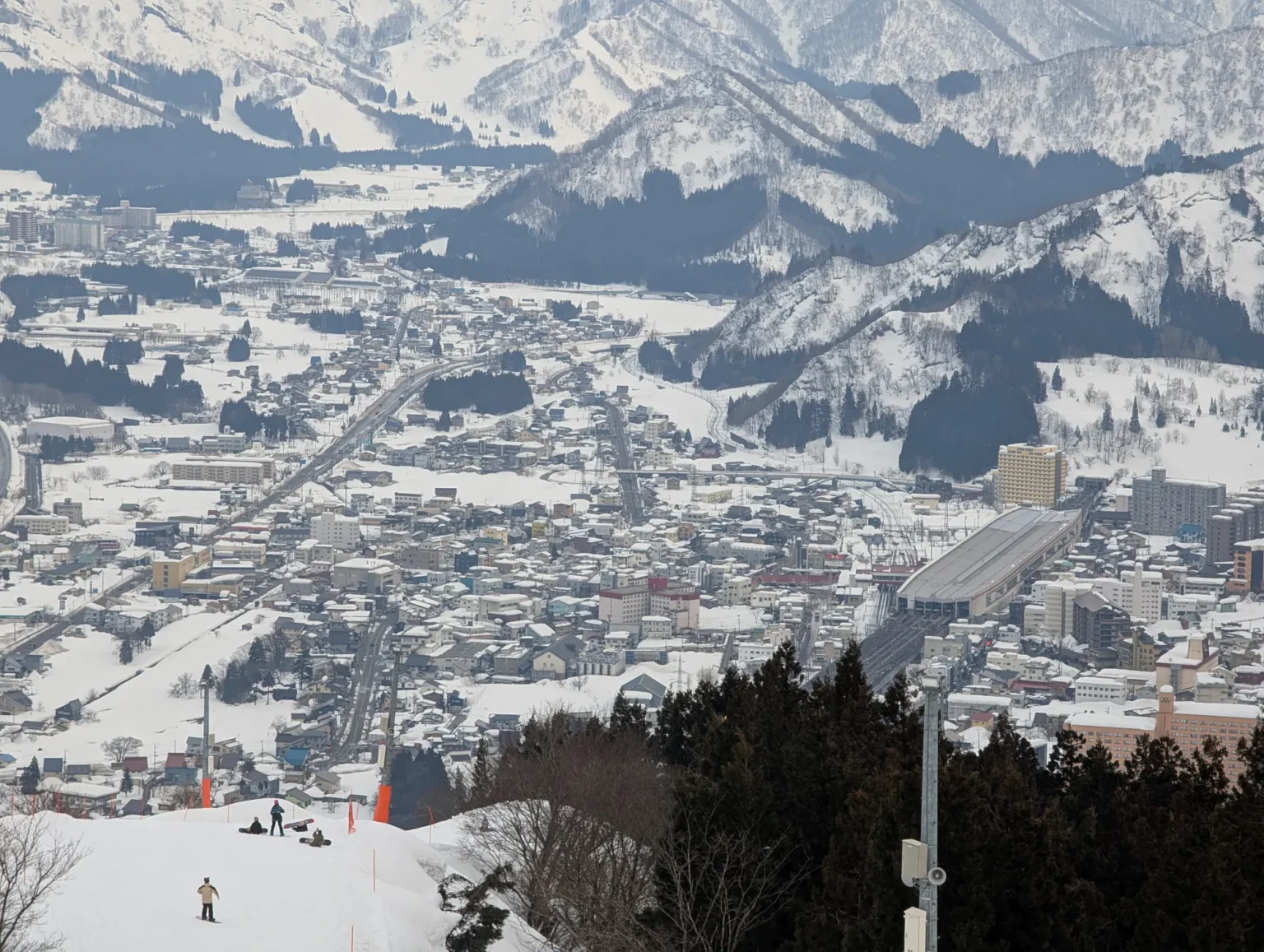 The view from GALA Yuzawa's slopes down over the snow-covered town of Yuzawa, with the long roofed platforms of Echigo-Yuzawa Station visible among the buildings and mountains rising behind.