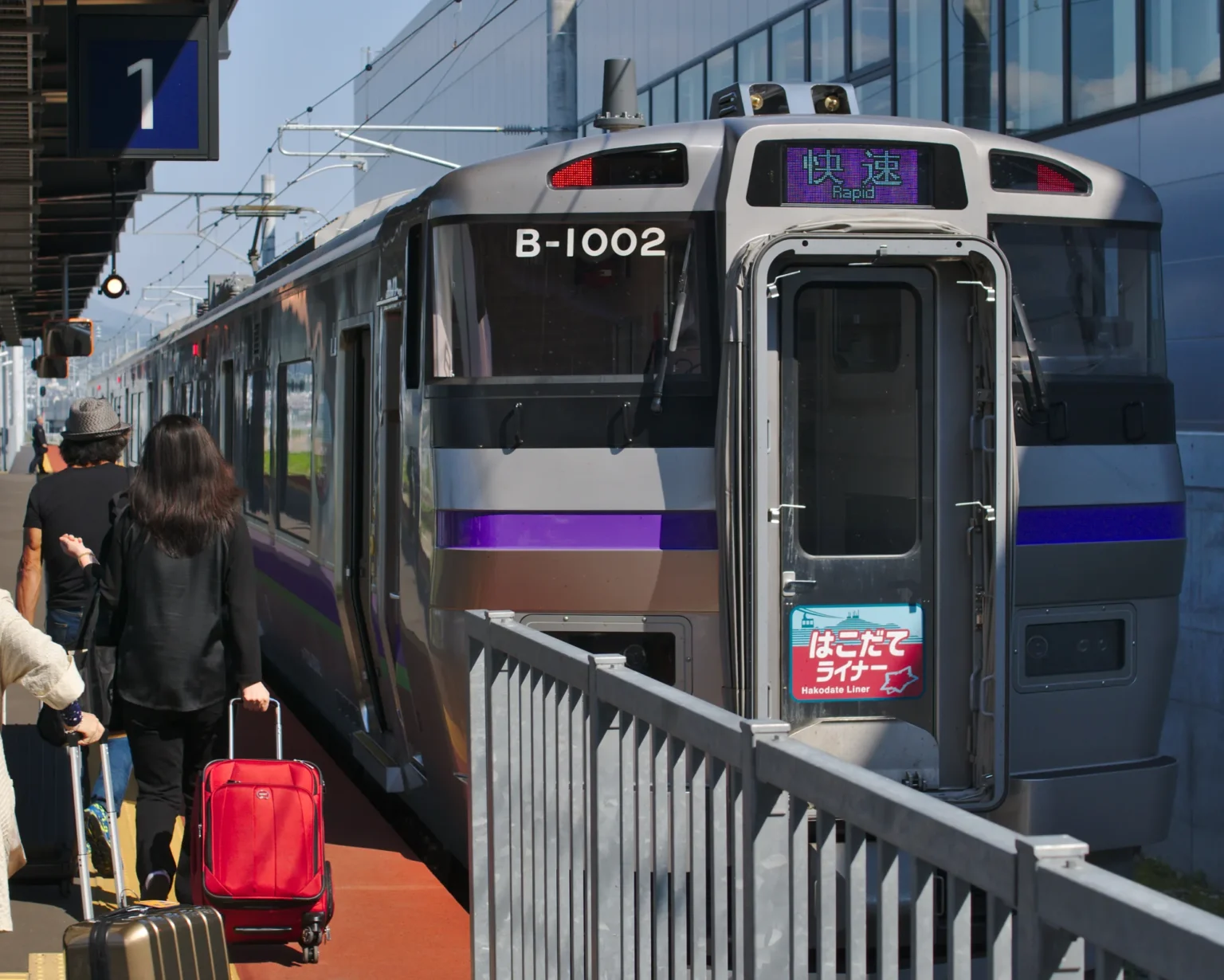 A silver 733 series Hakodate Liner train with a purple stripe and a Rapid destination sign waits at a Shin-Hakodate-Hokuto Station platform as passengers with suitcases walk beside it.