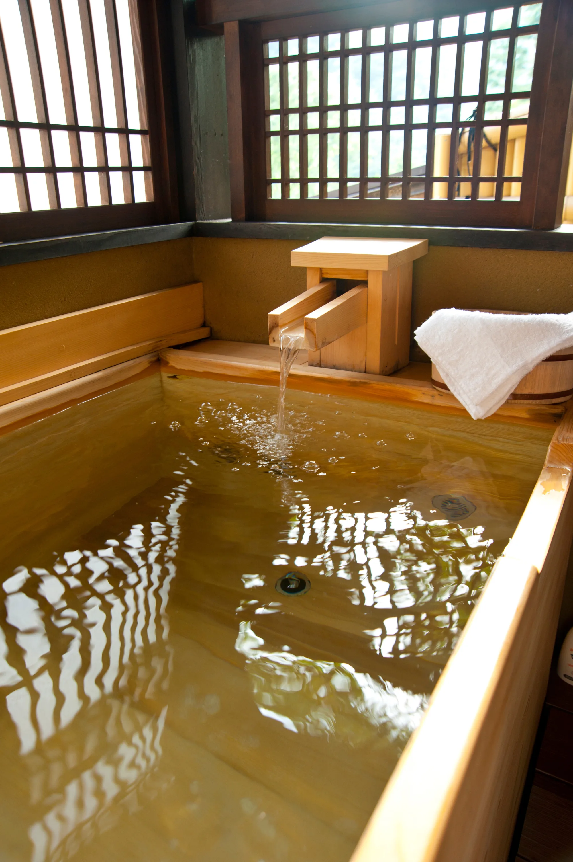 Hot spring water pouring from a wooden spout into a wooden bathtub at a Hakone onsen, with a folded white towel on the rim and latticed windows looking out to greenery behind