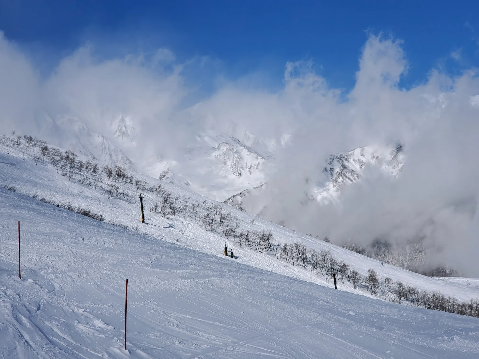 An upper piste at Happo-one: a broad snow slope with thin marker poles and scattered bare trees in the foreground, and cloud pouring over the snow-covered ridge of the Northern Alps behind it.