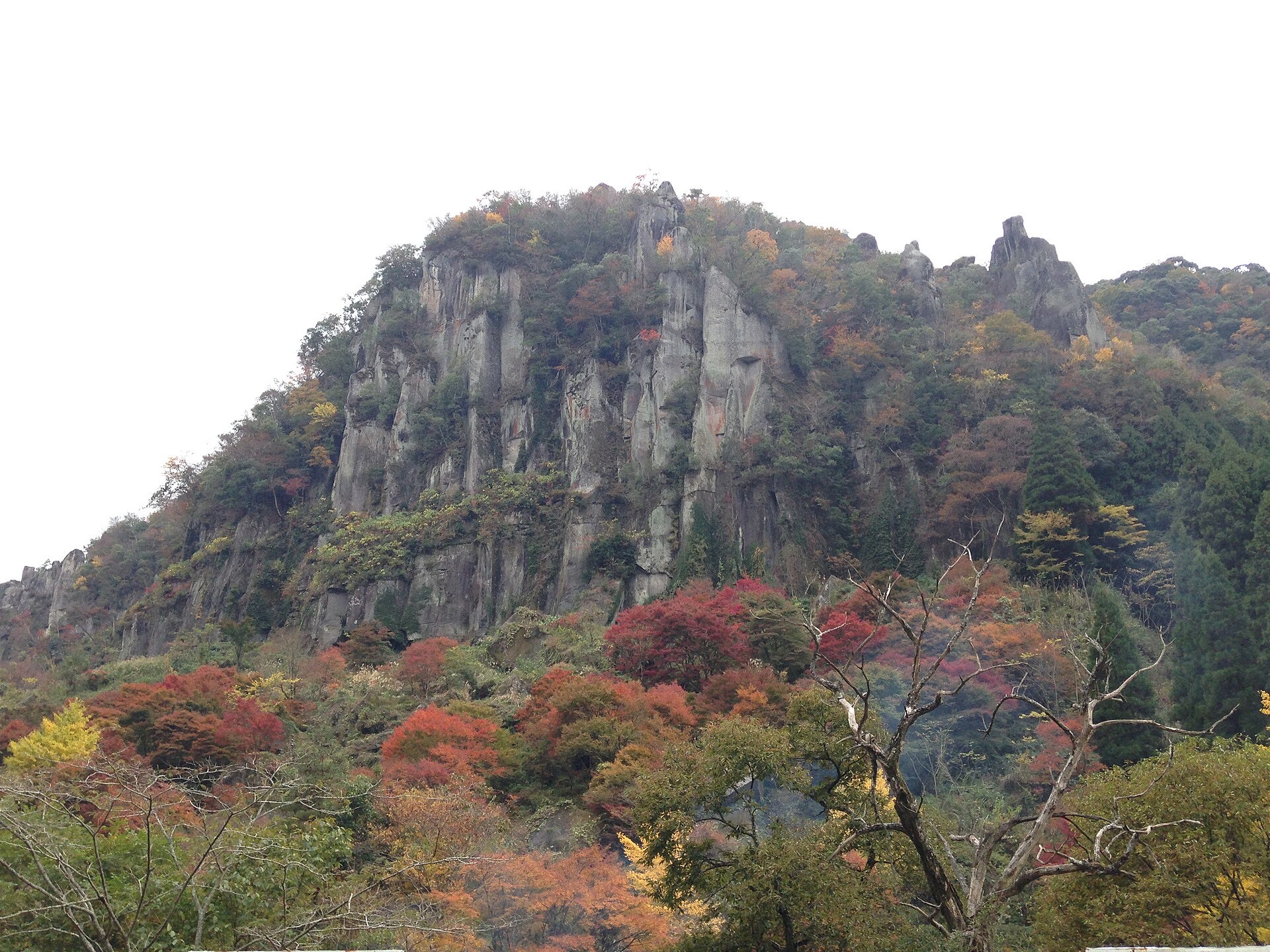 The eroded rock pinnacles of Hitome-Hakkei in Shin-Yabakei gorge, Oita, rising above a slope of red, orange and yellow autumn trees on an overcast November day
