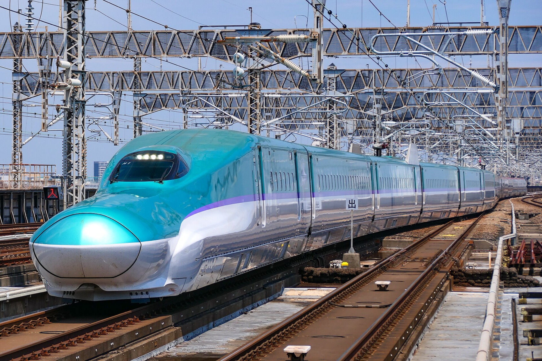 A teal and silver H5 series Hokkaido Shinkansen train with its long aerodynamic nose passing under steel gantries on elevated shinkansen tracks in bright daylight.