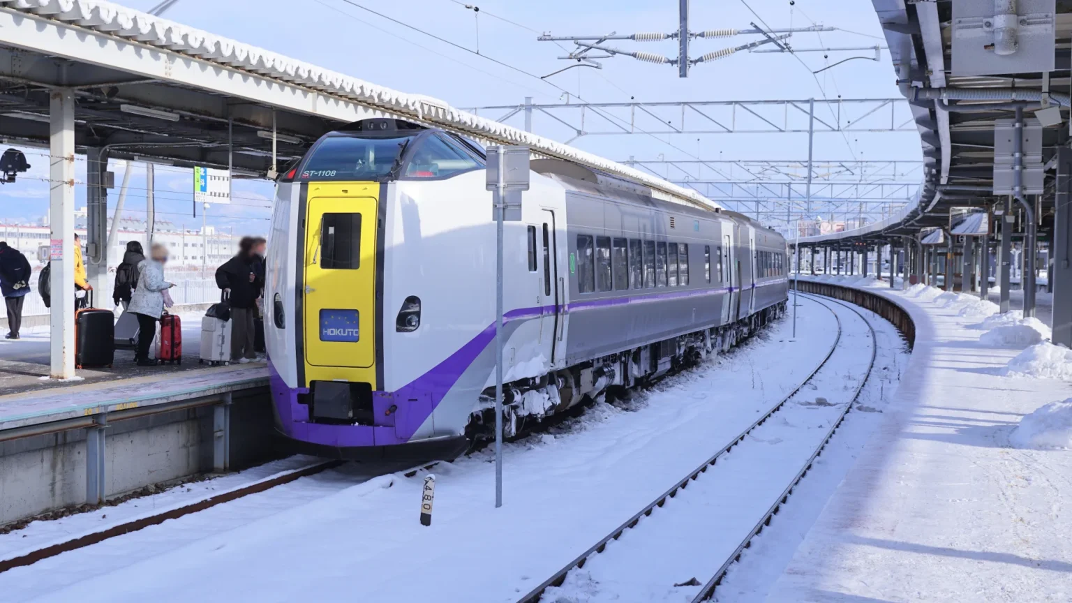 A white KiHa 261 series Hokuto limited express with a yellow front door and purple trim waits at a snow-covered platform of Hakodate Station while passengers with suitcases gather to board.