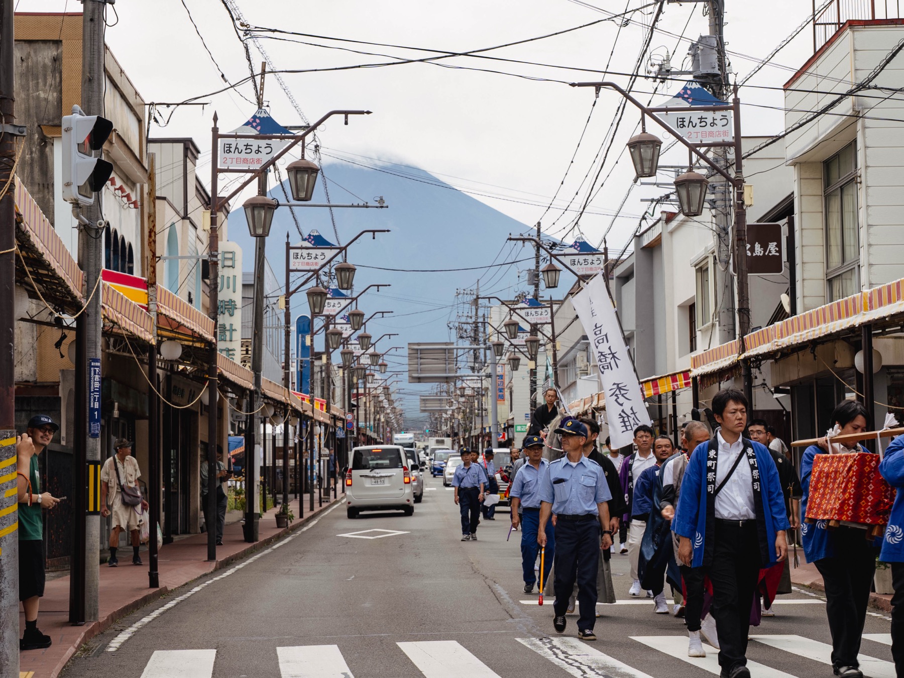 Festival participants in blue happi coats and police officers walk down the middle of Honcho-dori shopping street in Fujiyoshida with cloud-capped Mount Fuji filling the end of the street
