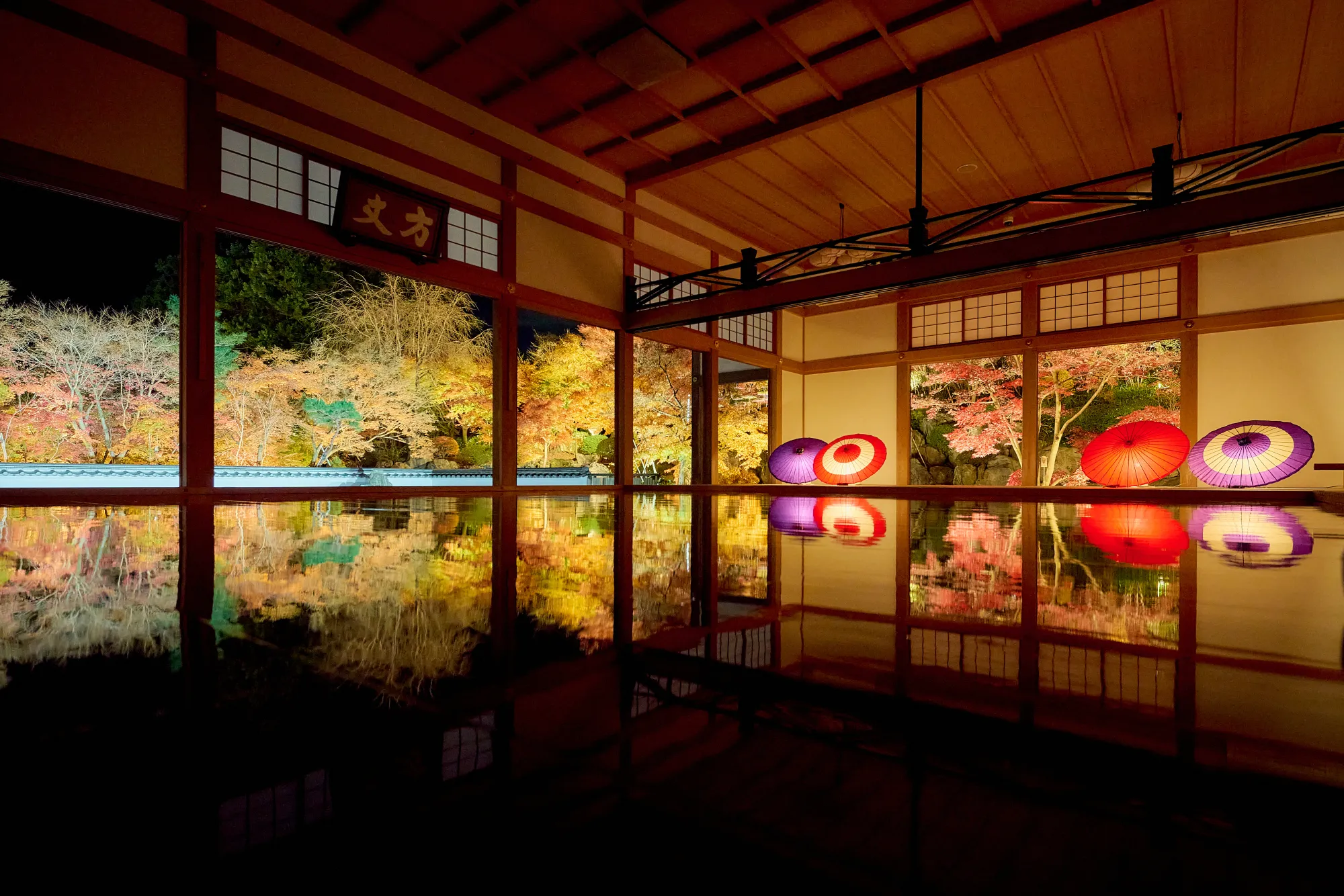 The same hall after dark during the November illumination: the garden outside is floodlit in gold, orange and red against a black sky, four paper parasols glow red and purple along the sill, and the whole scene repeats inverted in the polished floor.