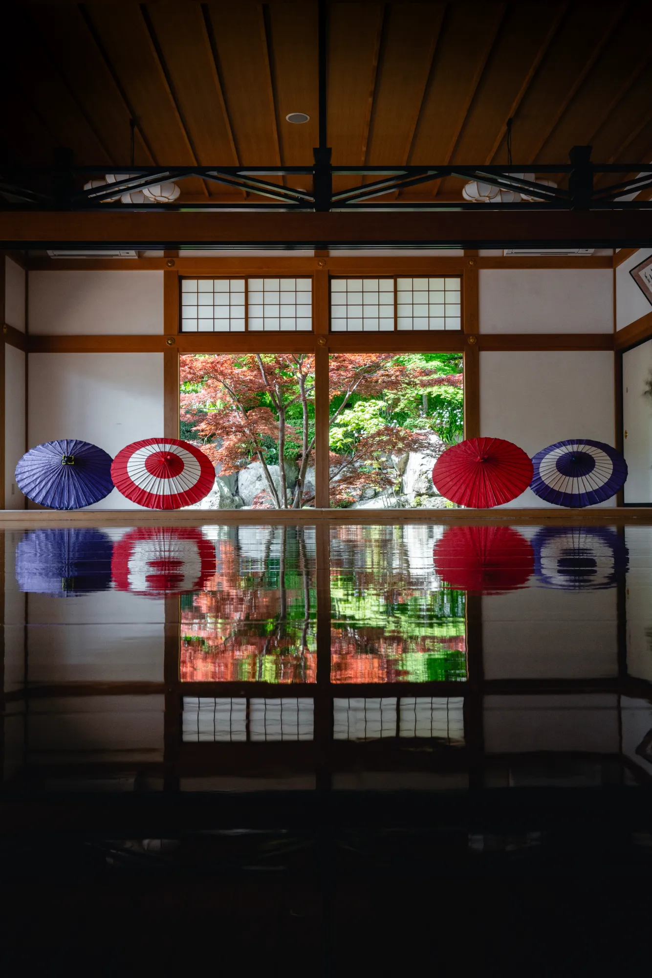 The same hall during the spring opening: a dim interior with one bright bay looking out on green and red maples over rocks, four paper parasols along the sill, and a reflection in the black floor almost as strong as the view itself.