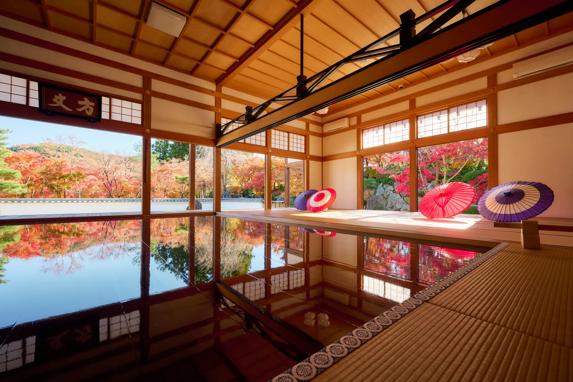 The main hall at Hotokuji in autumn: window bays open on two sides onto red and orange maples and a rock garden, three paper parasols stand on the raised tatami, and the broad dark floor in the foreground reflects the maples, the window frames and the ceiling beams upside down.