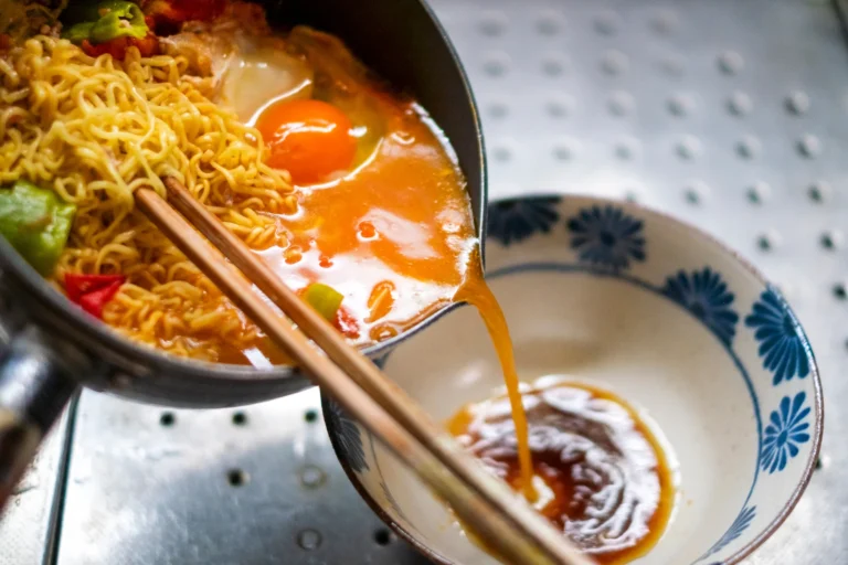 Instant ramen noodles with a soft egg being ladled from a metal pot into a blue-and-white Japanese porcelain bowl, with chopsticks resting across the rim and broth pouring from the ladle