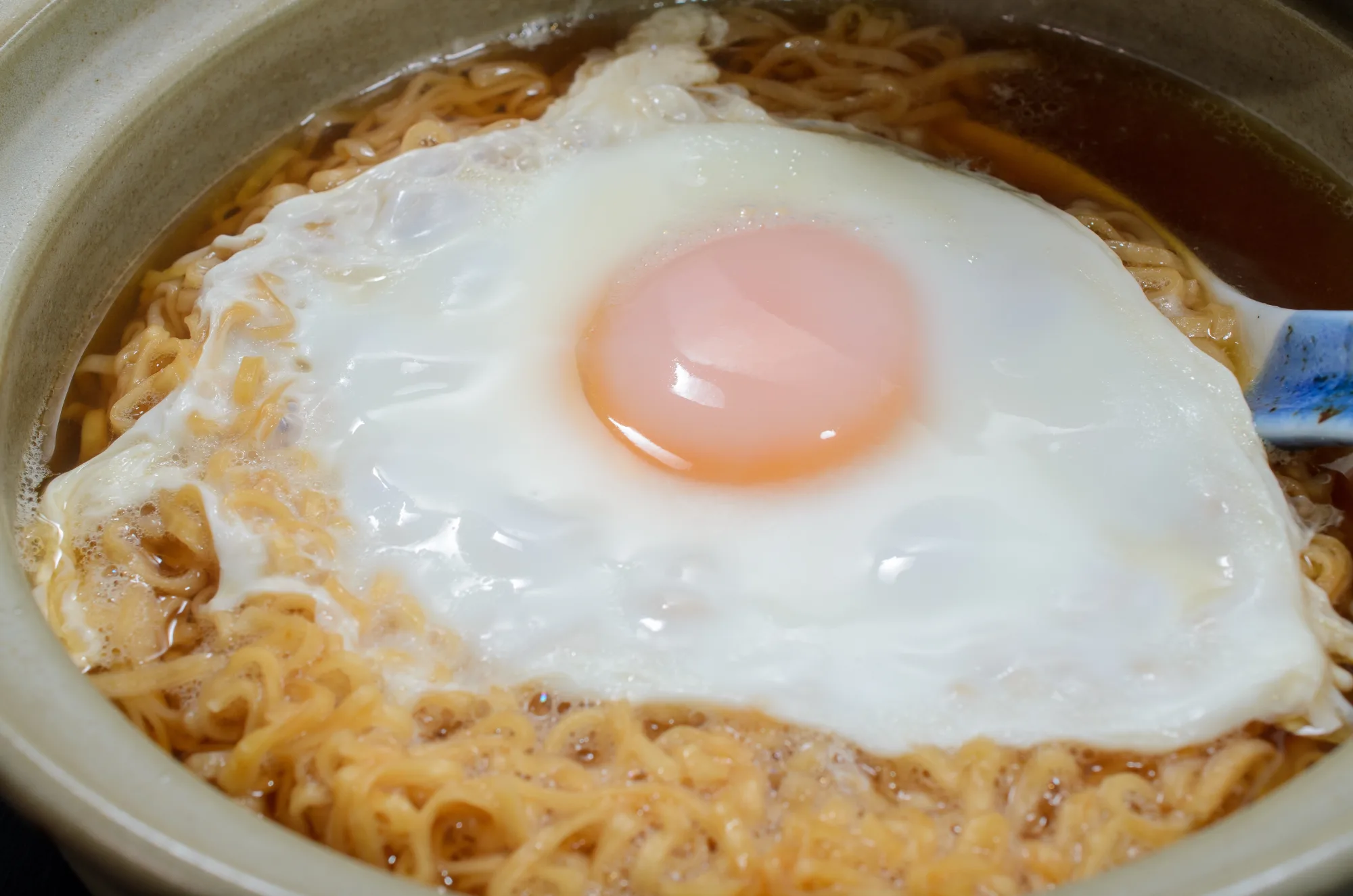 A fried egg with a bright orange yolk sitting on top of instant ramen noodles in a dark clay donabe pot, with the broth visible around the edges of the noodles