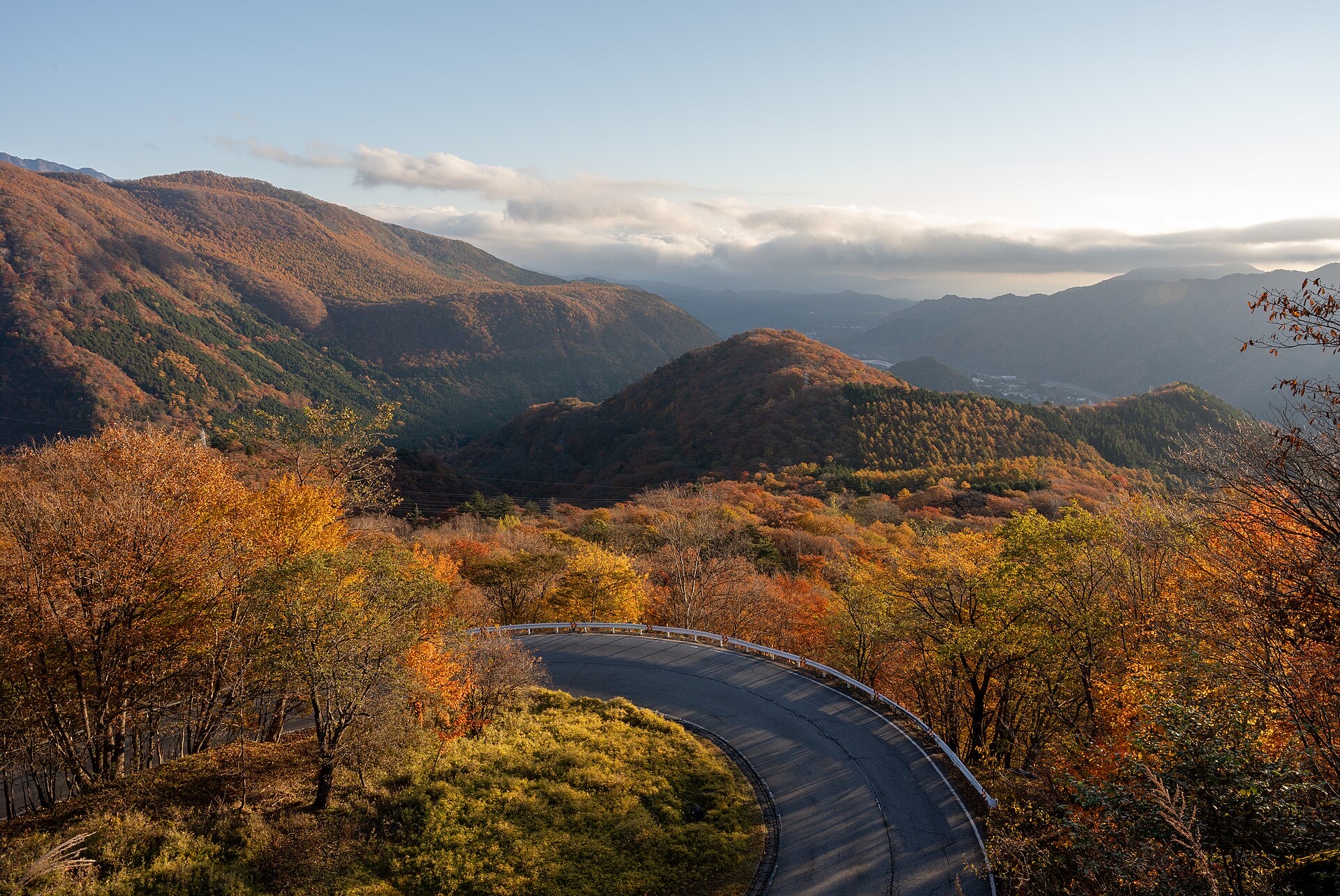 A hairpin curve of the Irohazaka winding road in Nikko in low autumn light, with orange and gold forest falling away to hazy mountain ridges beyond