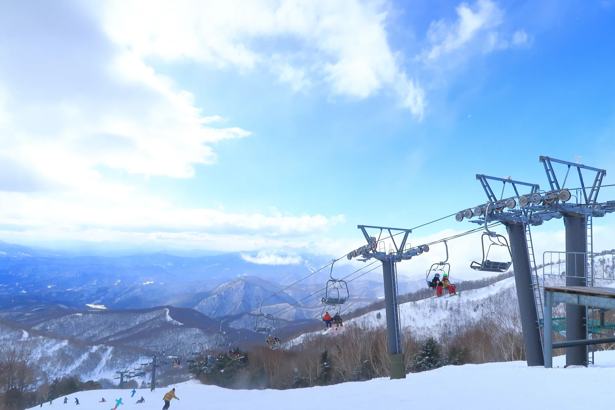 A two-person chairlift carrying skiers up a Japanese ski resort on a bright winter day, with groomed piste below and forested mountain ridges receding into the distance under scattered cloud