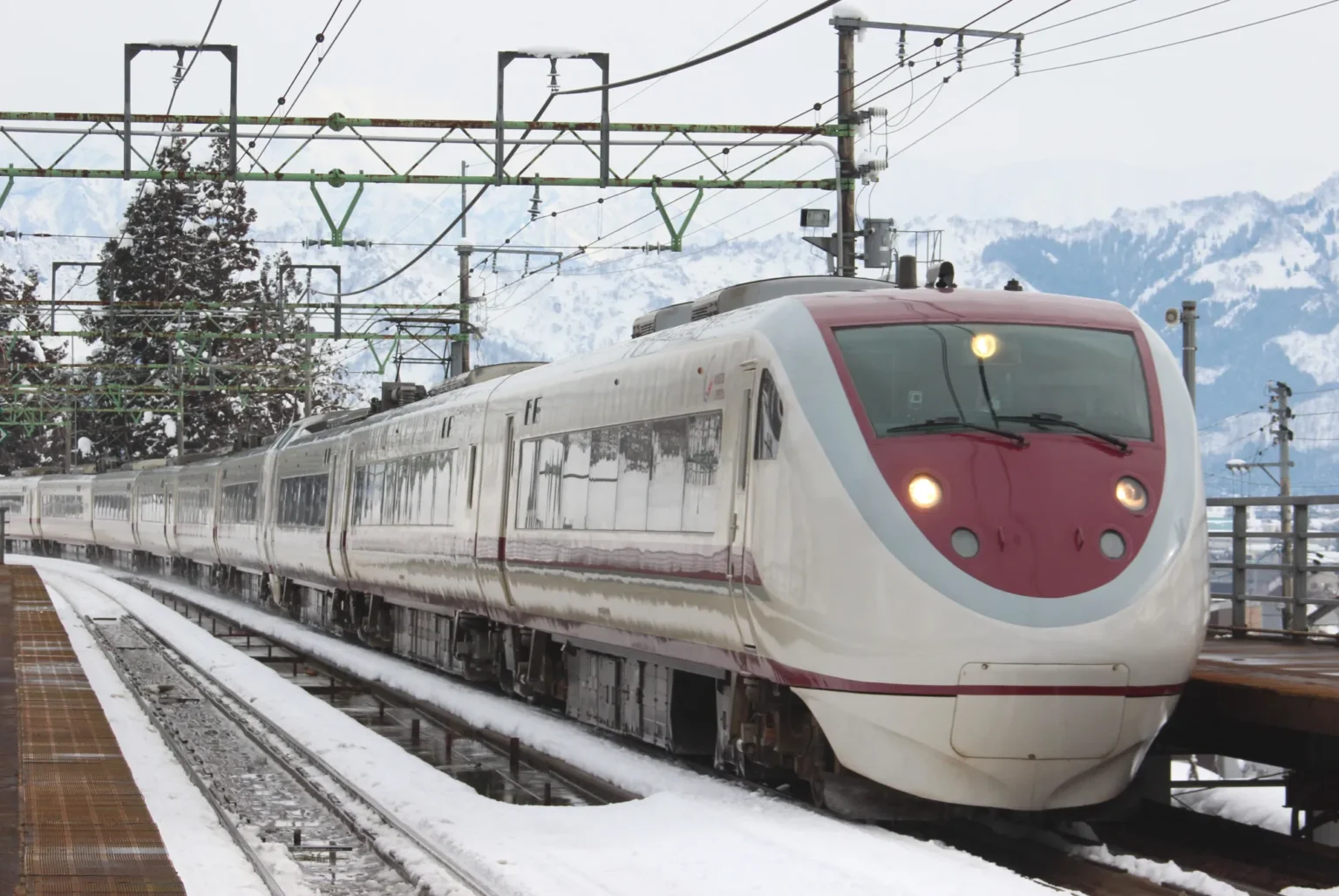 A white-and-crimson limited express train stopped at the snow-covered platform of Joetsu-Kokusai-Skijo-mae station, with snowy mountains behind.