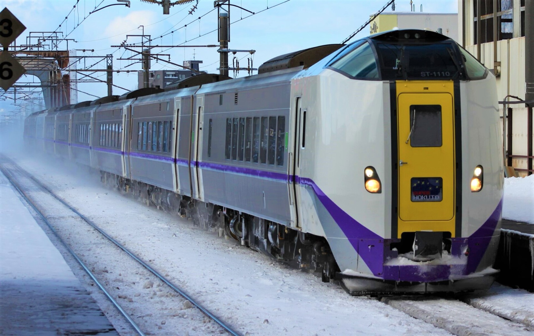 The Super Hokuto limited express with its distinctive yellow front door standing at a snow-covered platform, powder snow swirling around the carriages on a clear winter morning.