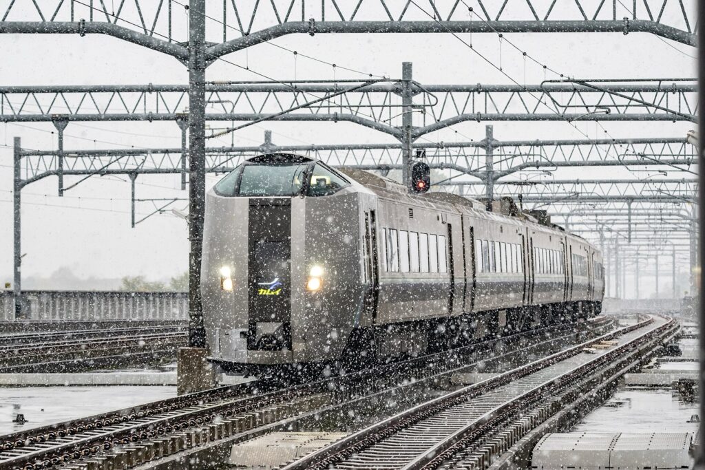 A silver JR Hokkaido 789 series Kamui limited express train approaching through heavy falling snow under overhead line gantries, headlights glowing against the grey winter sky.