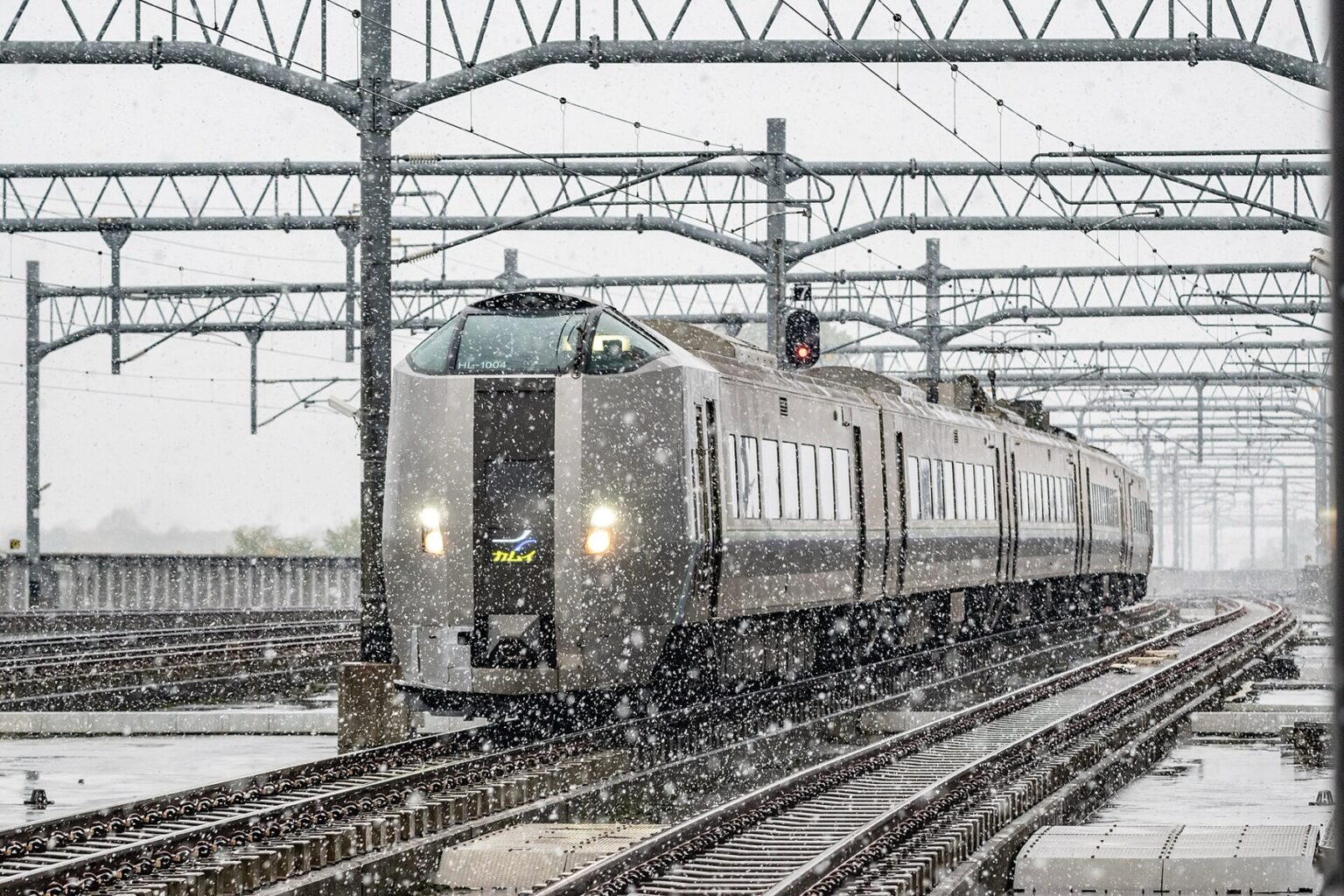 A silver JR Hokkaido 789 series Kamui limited express train approaching through heavy falling snow under overhead line gantries, headlights glowing against the grey winter sky.