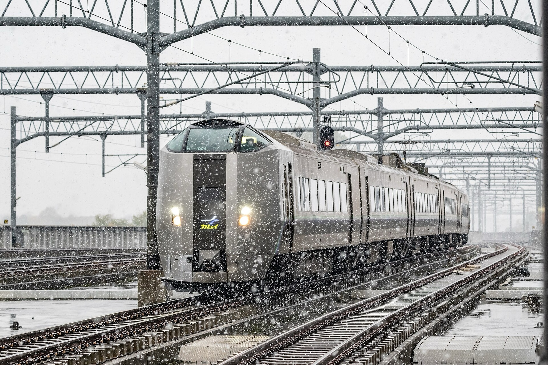A silver JR Hokkaido 789 series Kamui limited express train approaching through heavy falling snow under overhead line gantries, headlights glowing against the grey winter sky.