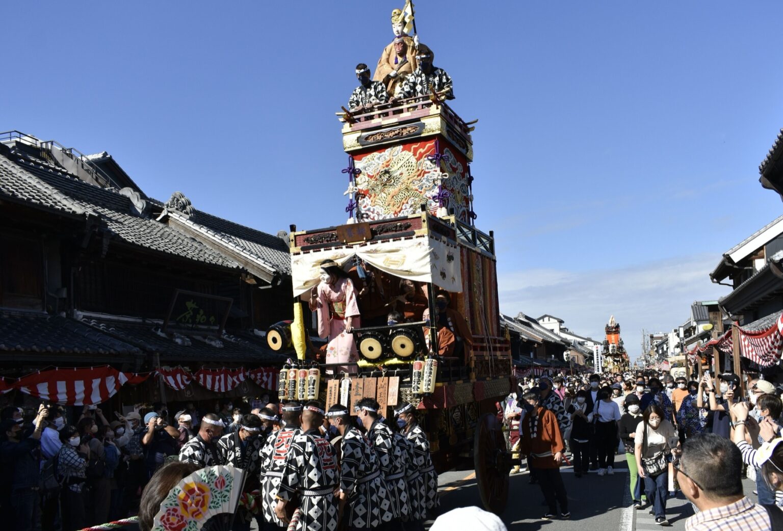 A gilded Kawagoe Festival float pulled along Ichibangai street by teams in black-and-white patterned happi coats, a doll figure crowning the top tier and traditional warehouse-style merchant buildings lining the road under a blue sky.