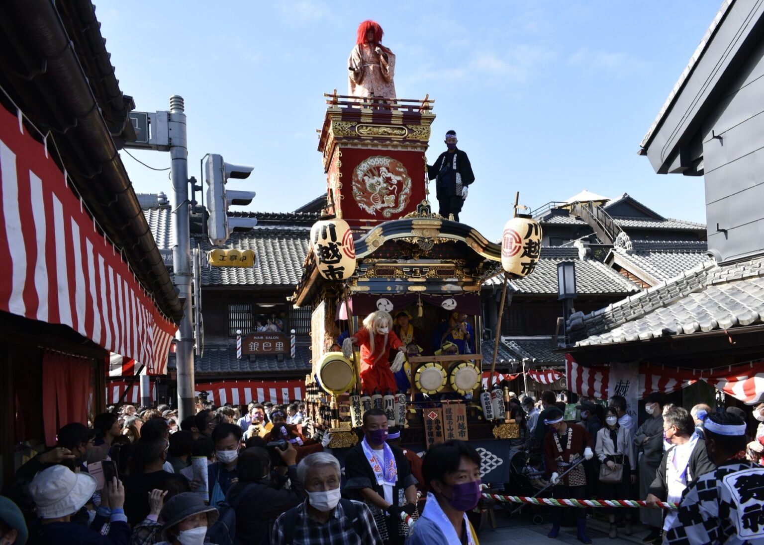 A tall Kawagoe Festival float with a red-robed doll figure on its upper tier rolling through the narrow kurazukuri merchant street, red and white festival bunting on both sides and a dense daytime crowd below.