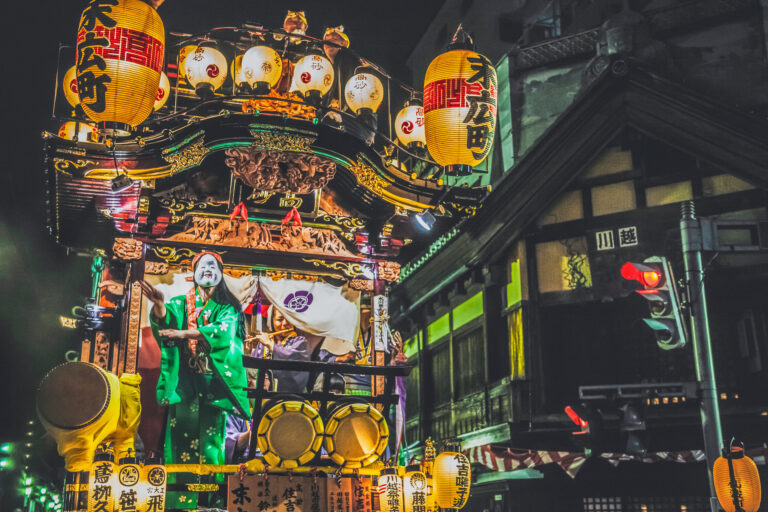 A Kawagoe Festival dashi float at night, its tiered black-and-gold body hung with dozens of glowing white lanterns reading Suehiro-cho, a masked dancer in green performing on the stage as the float faces a dark wooden merchant building.