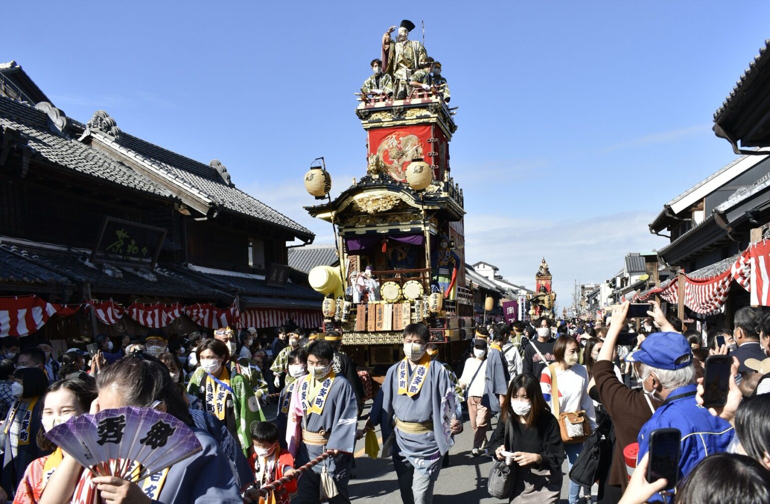 Two Kawagoe Festival floats facing each other down the length of the kurazukuri street, musicians and masked performers on their stages, with festival-goers in blue happi coats and spectators filling the road between them.
