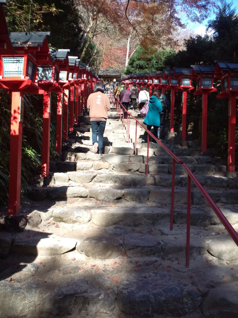 The stone staircase at Kifune Shrine in Kyoto lined on both sides with tall vermilion kasuga lanterns, visitors climbing between them and autumn-tinted maples on the hillside above.