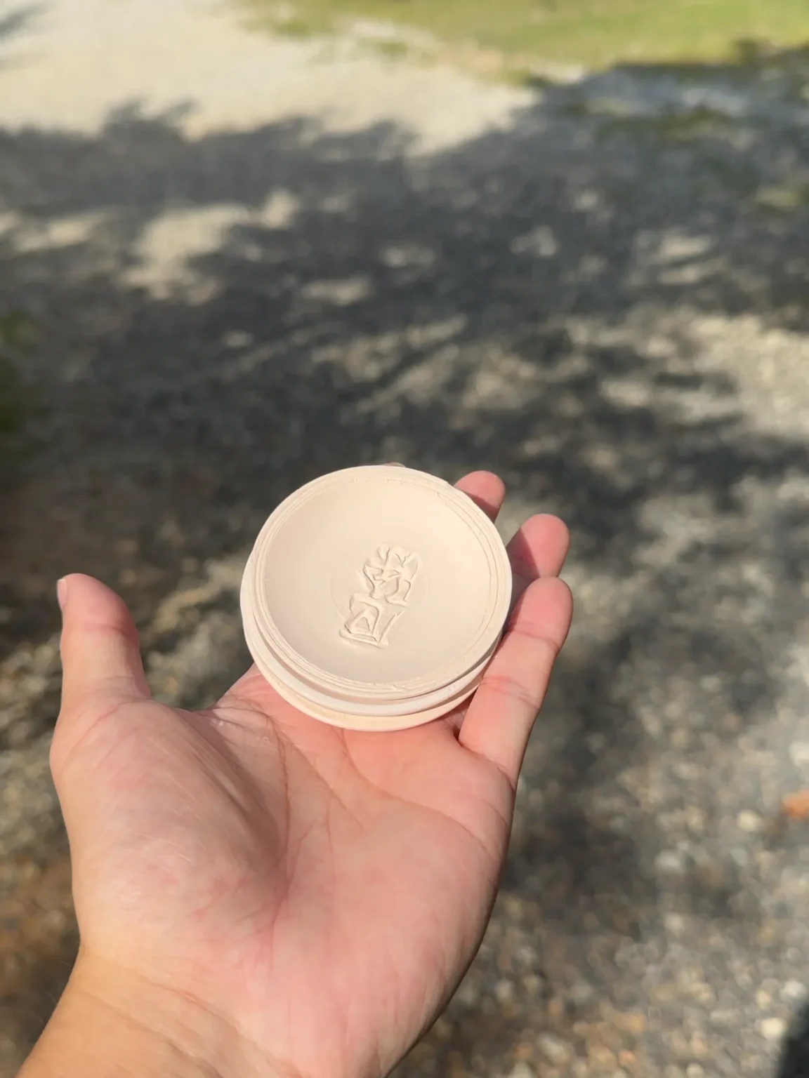 A hand holding a small stack of unglazed kawarake clay discs, the top one stamped with a character, over a gravel path at the Daishi-yama summit above Kinosaki Onsen