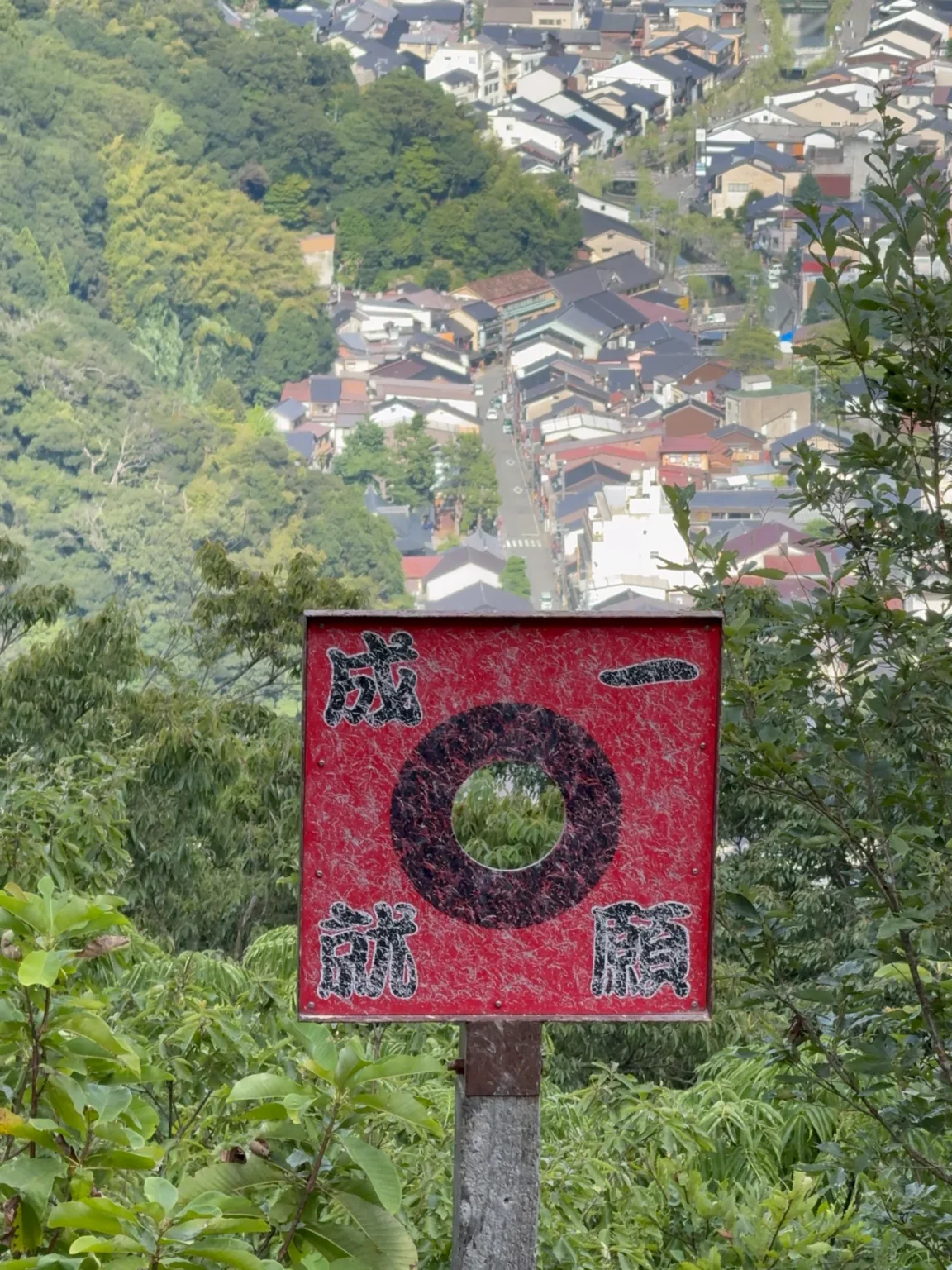 A red square target with a black ring around a hole, painted with the four characters for one wish fulfilled, on a post on the wooded slope below the kawarake-throwing spot on Daishi-yama, with the rooftops and main street of Kinosaki Onsen far below