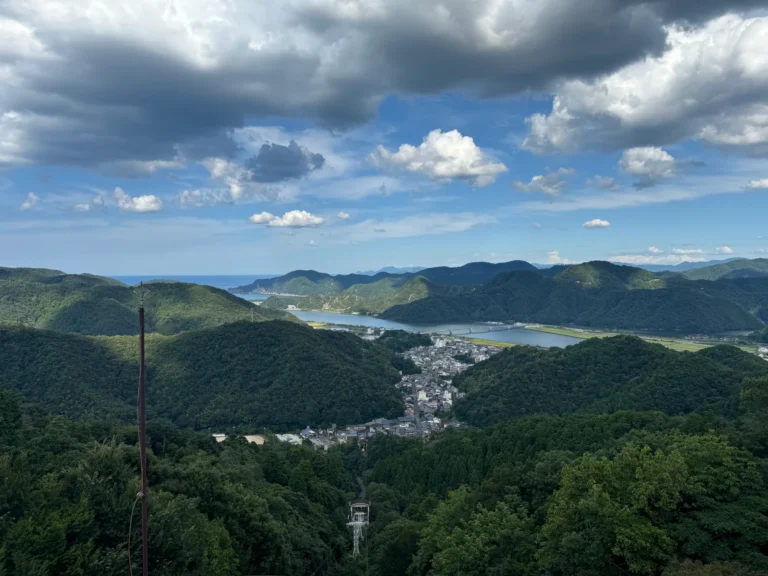 Kinosaki Onsen seen from the Daishiyama summit: the hot-spring town fills a wooded valley, with the Maruyama River, a long bridge and the Sea of Japan on the horizon, and a ropeway tower in the forest below